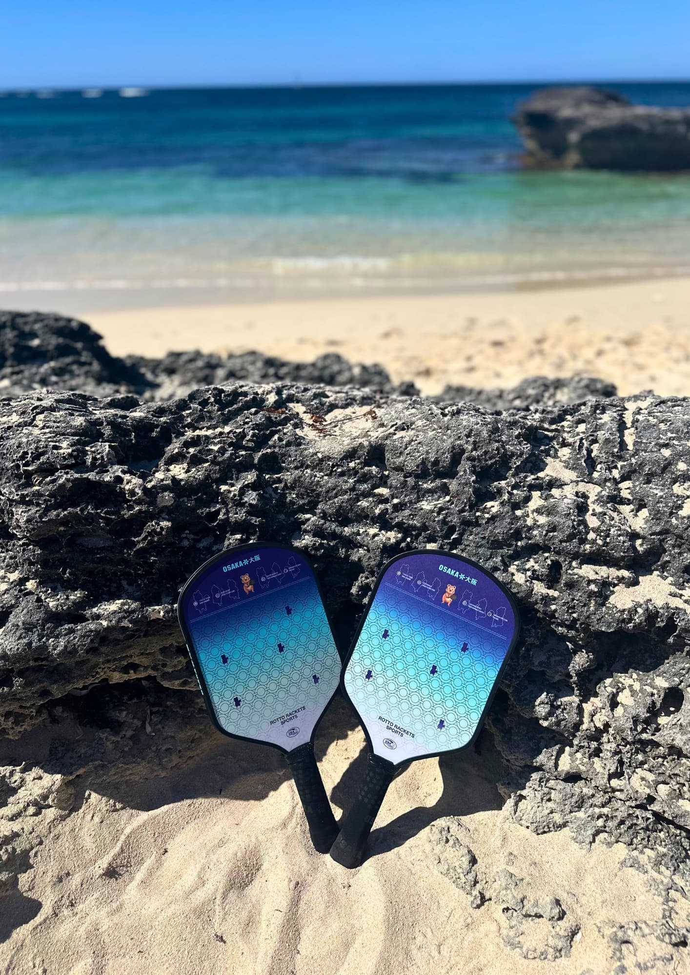 Two pickleball paddles lying in sand on beach in front of rocks and blue water
