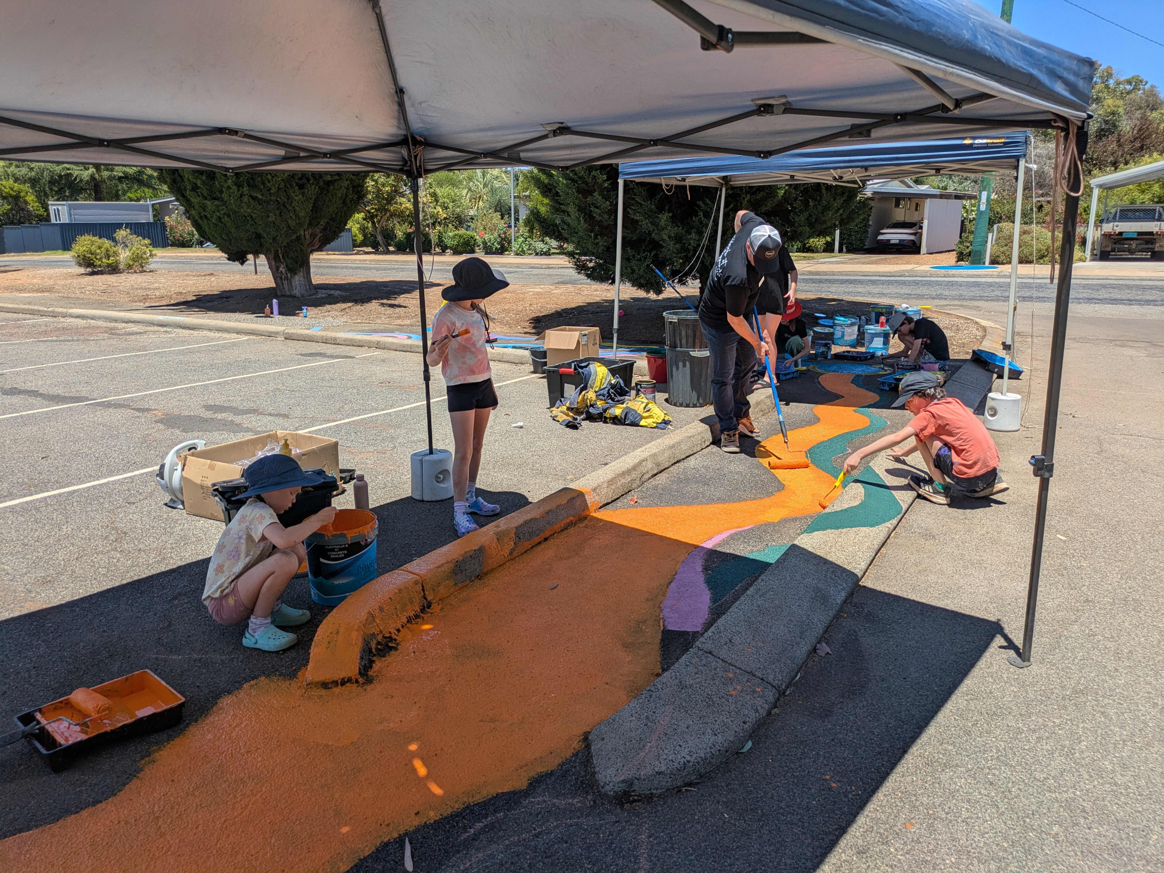 Children painting a colourful mural on concrete street under shade.