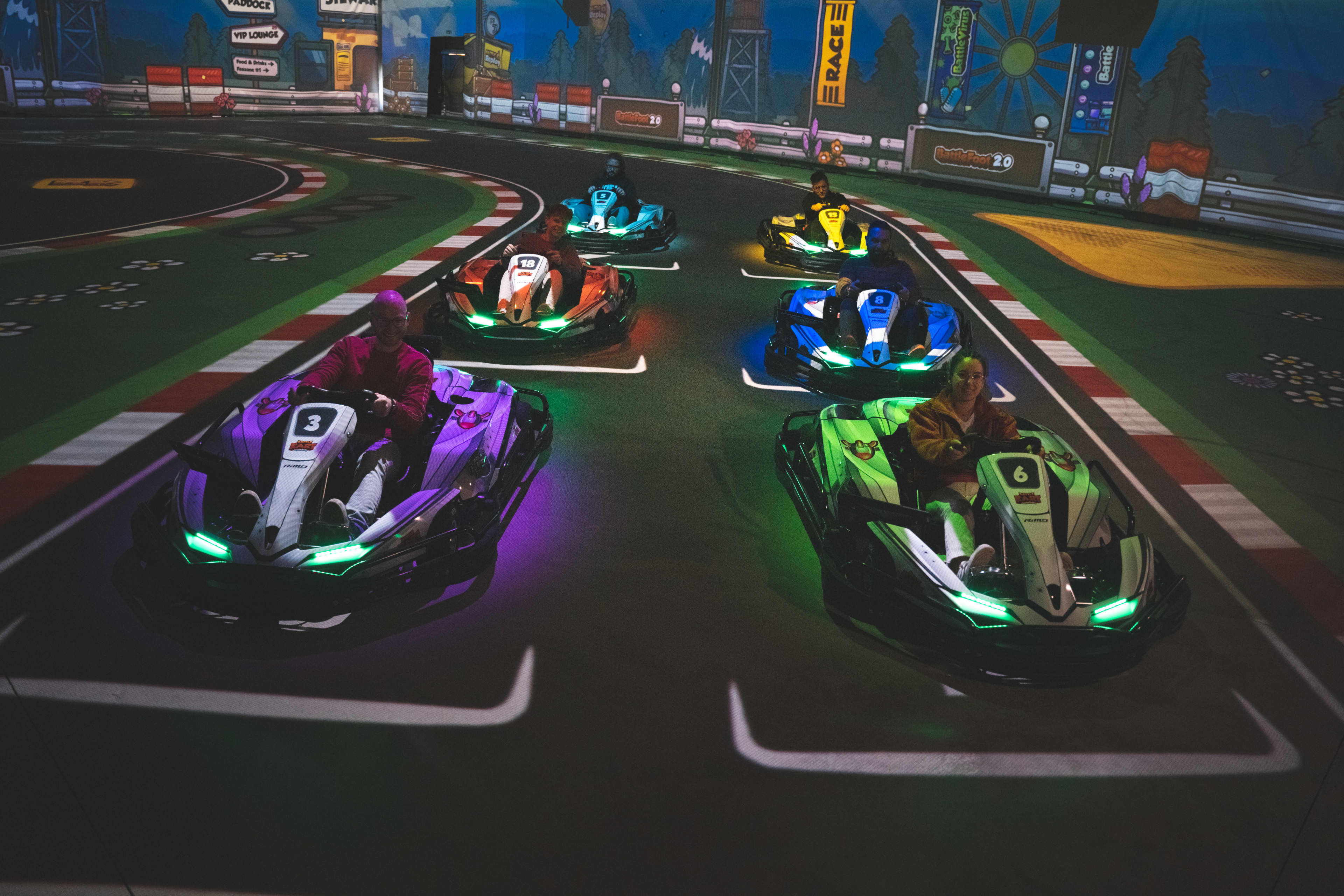 Six people sit in colorful electric go-karts lined up on an indoor racetrack, viewed from the front. The karts have glowing LED lights and visible numbers, positioned on starting grid lines with a curving track, barriers, and race-themed decorations in the background.