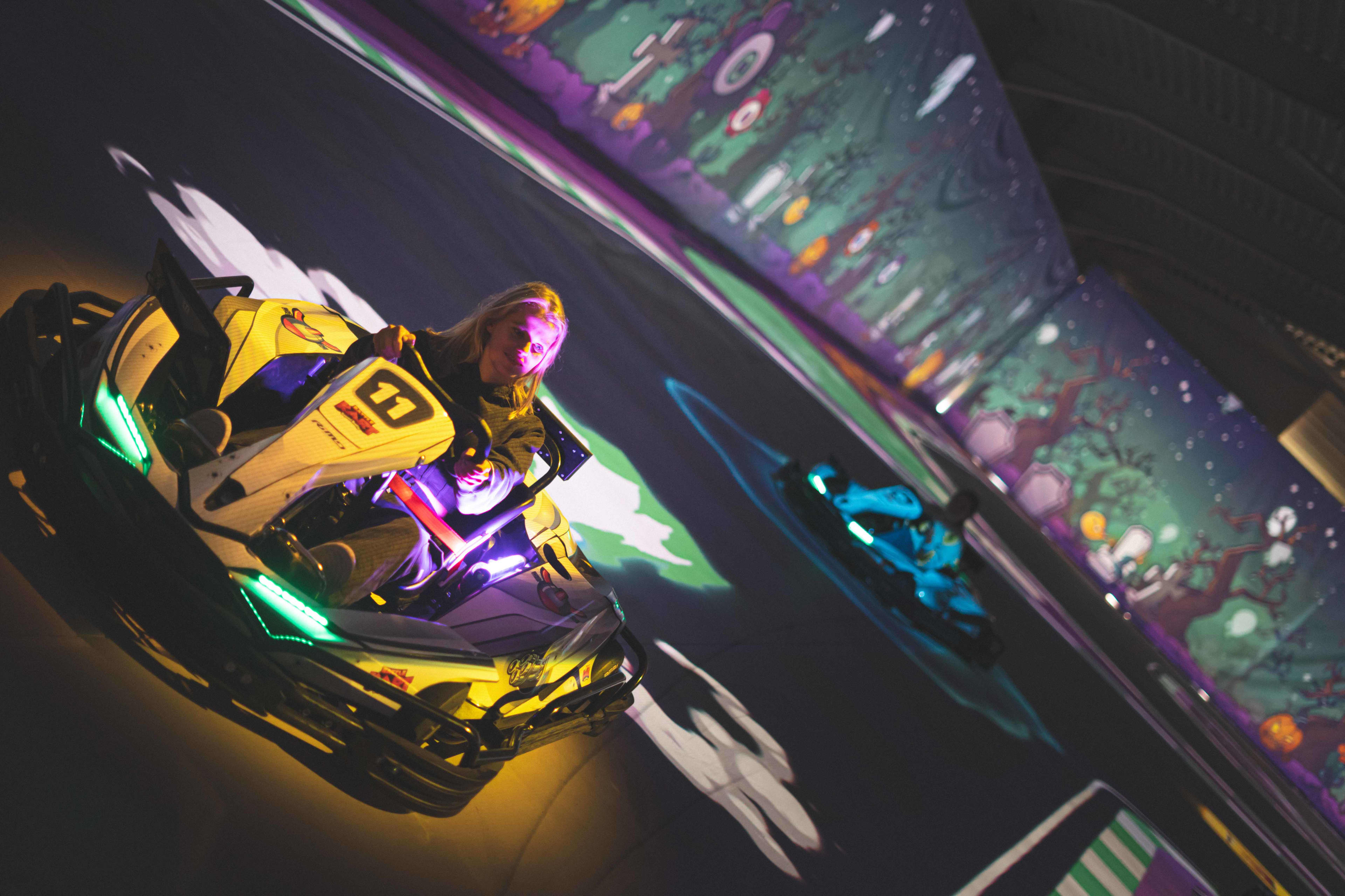 A driver in a yellow electric go-kart with glowing green and purple lights races through an indoor track with projected, game-like graphics on the floor and walls. The kart is angled mid-turn, with another illuminated kart following behind. The dark arena is filled with colorful projections and neon lighting, creating a dynamic, futuristic racing atmosphere.