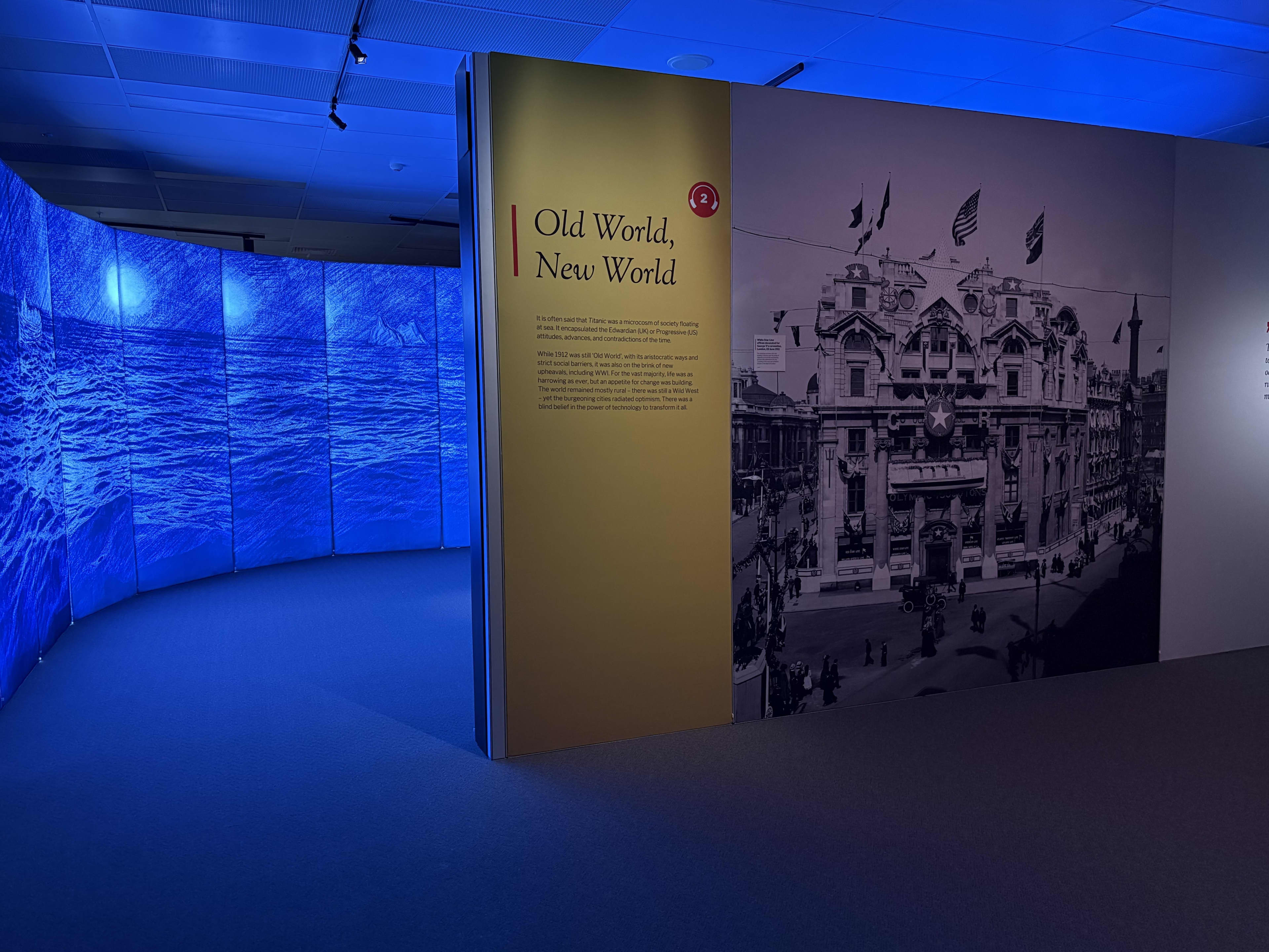 Museum gallery interior with blue lighting: on the left, a curved immersive display showing a stylized ocean scene; in the center, a tall yellow panel titled “Old World, New World”; on the right, a large black-and-white historical photograph of an ornate early-20th-century building decorated with flags and crowds below.
