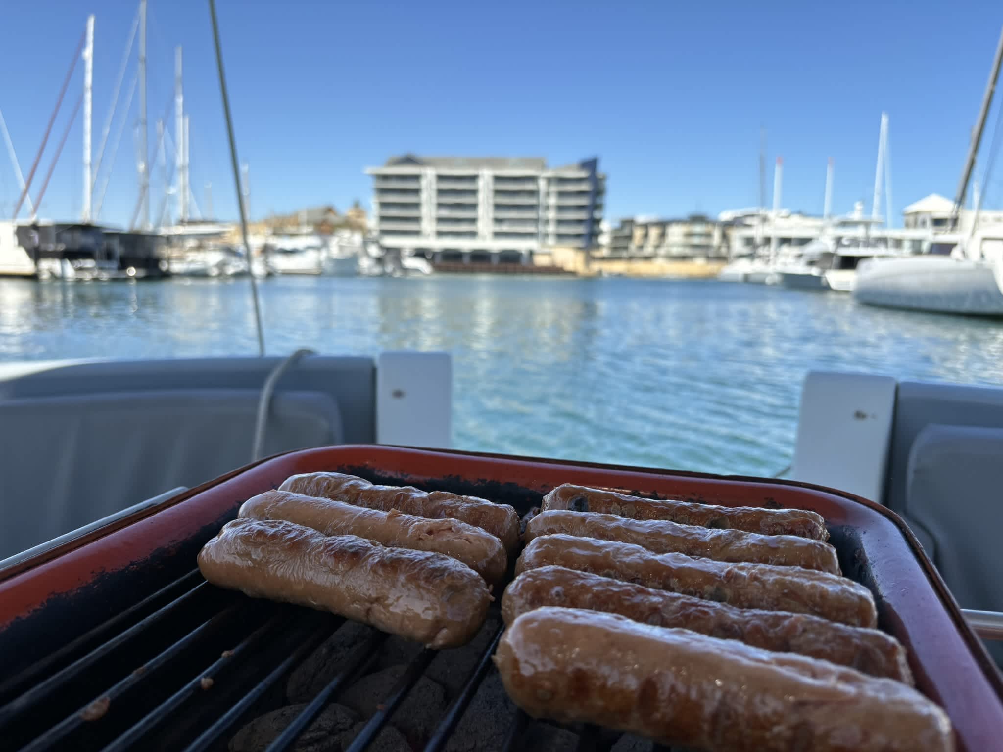 8 sausages sitting on a grill overlooking a blue marina filled with sail boats.