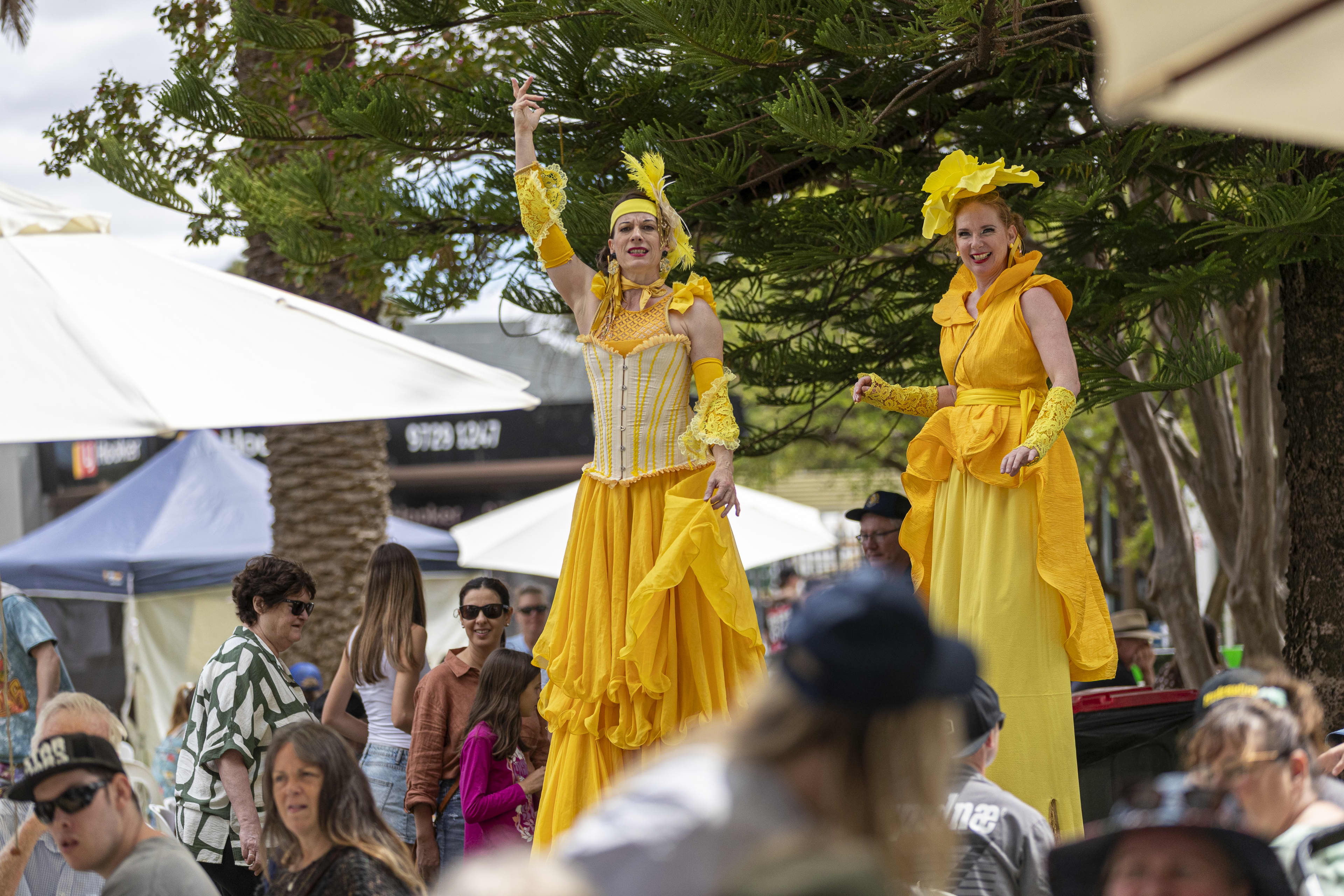 Two yellow performers on stilts standing amongst a crowd of people roaming the market stalls.