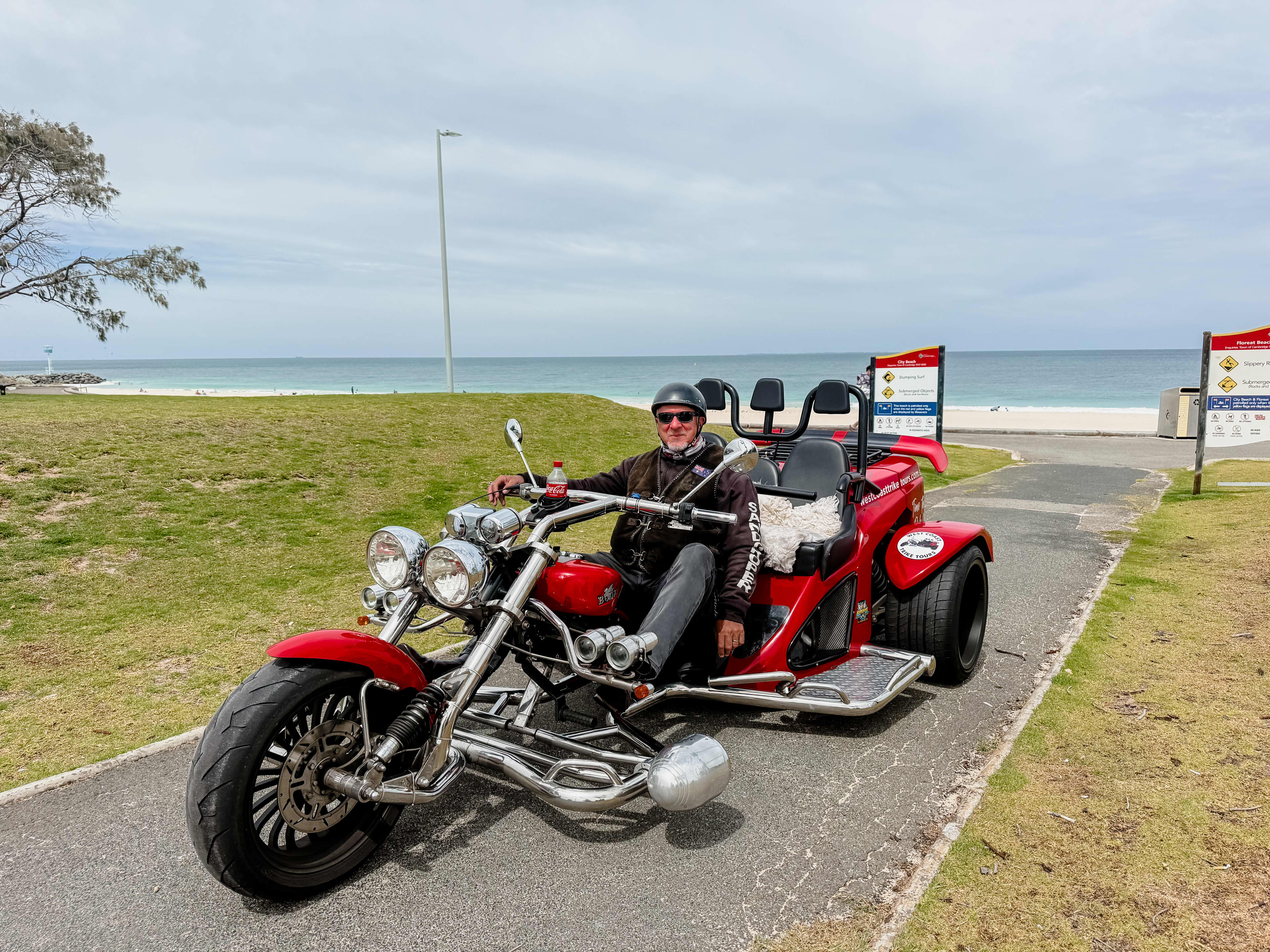 A person wearing a helmet and sunglasses sits on a red three-wheeled motorcycle parked on a paved path near a grassy beach area, with the ocean and cloudy sky in the background.