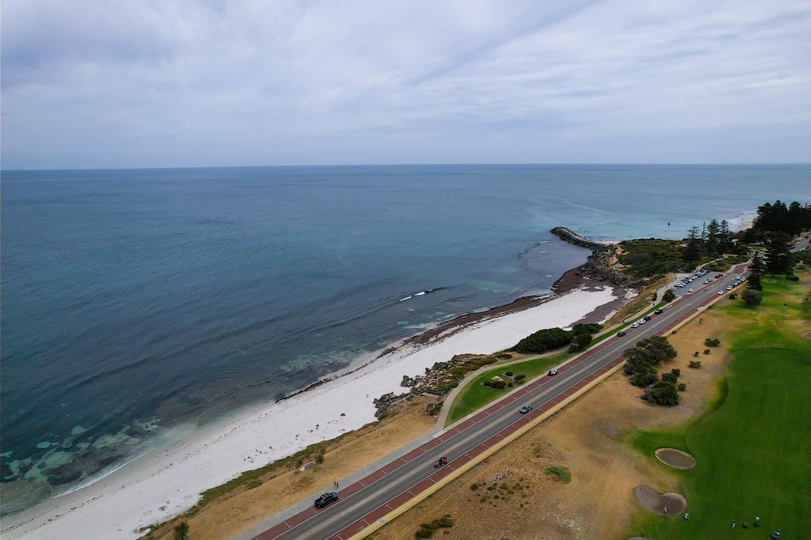 Aerial view of a coastal landscape showing a long road running parallel to a white sandy beach, with calm blue ocean on the left and grassy parkland on the right, scattered cars on the road, and a rocky headland extending into the water under a lightly overcast sky.
