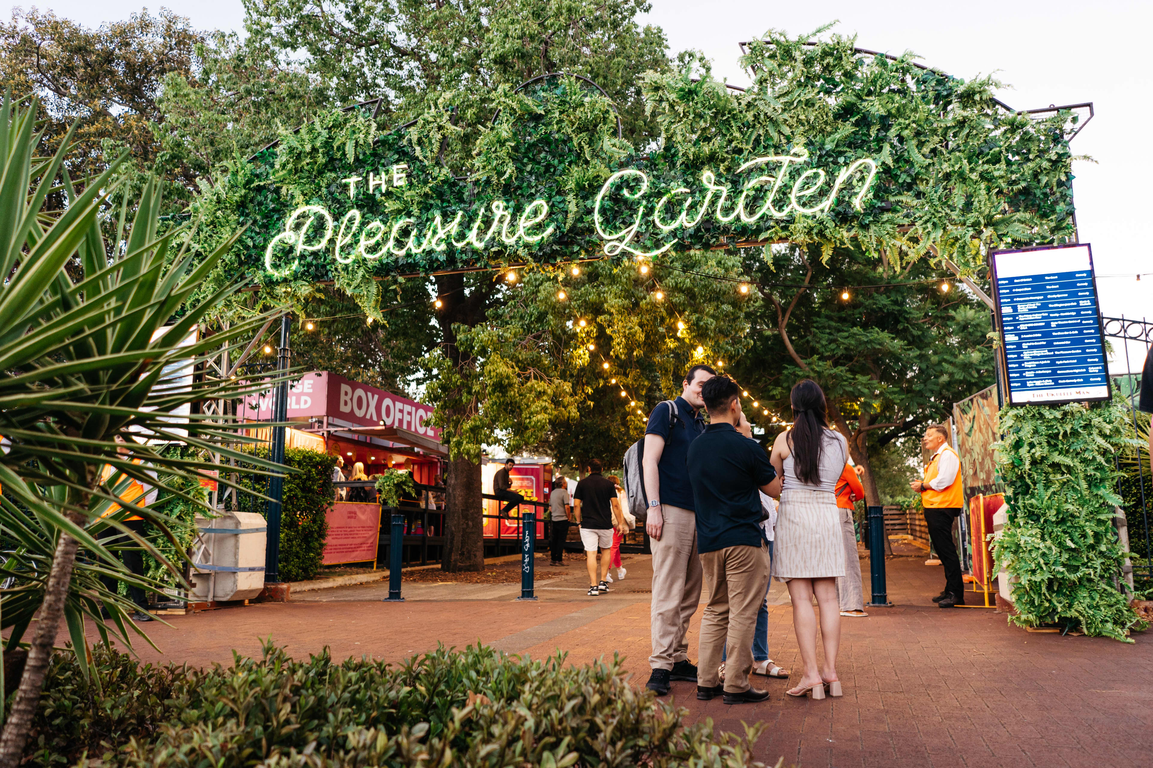 A lush green arch featuring a neon lit sign reading "The Pleasure Garden", with three people standing outside the front entrance.