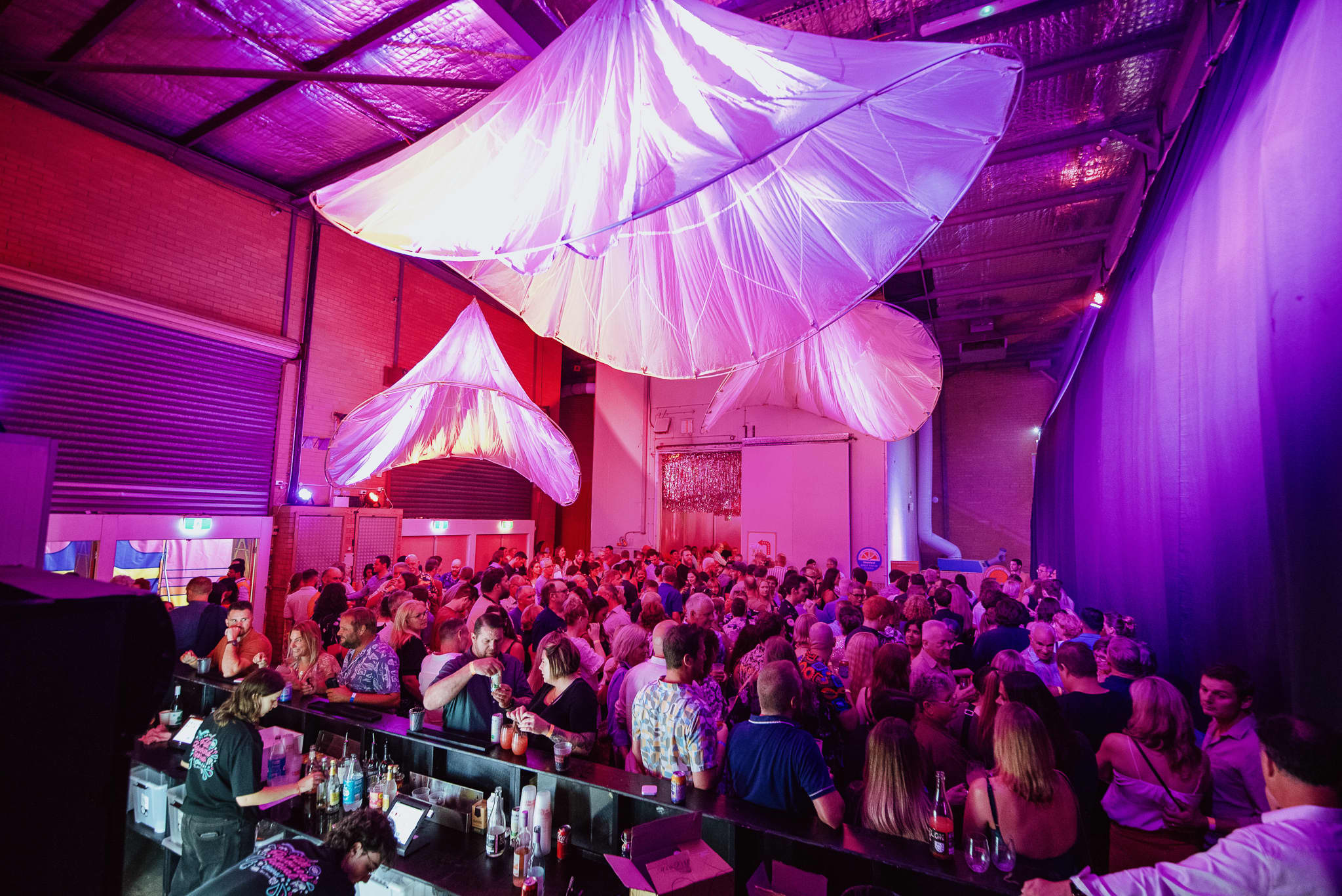 A crowded indoor event space with a long bar in the foreground and a large group of people socializing beneath dramatic purple and pink lighting, with translucent fabric canopies suspended from the ceiling creating a festive, immersive atmosphere.