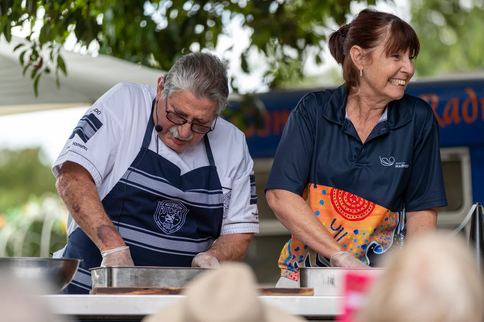Two adults prepare food at an outdoor event. An older man wearing glasses, a headset microphone, and a butcher’s apron focuses on mixing ingredients in a metal bowl, while a woman beside him in a navy shirt and colorful apron smiles and looks to the side. They stand behind a table with bowls and cutting boards, with trees and a blurred audience in the background.