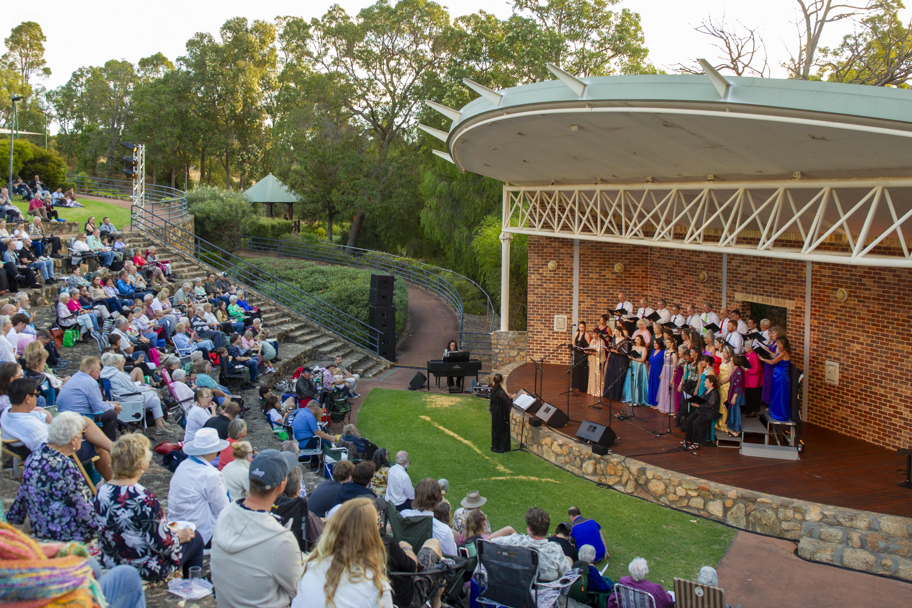 A group of singers performing on an outdoor stage, with a large lawn amphitheatre of adults watching the performance.