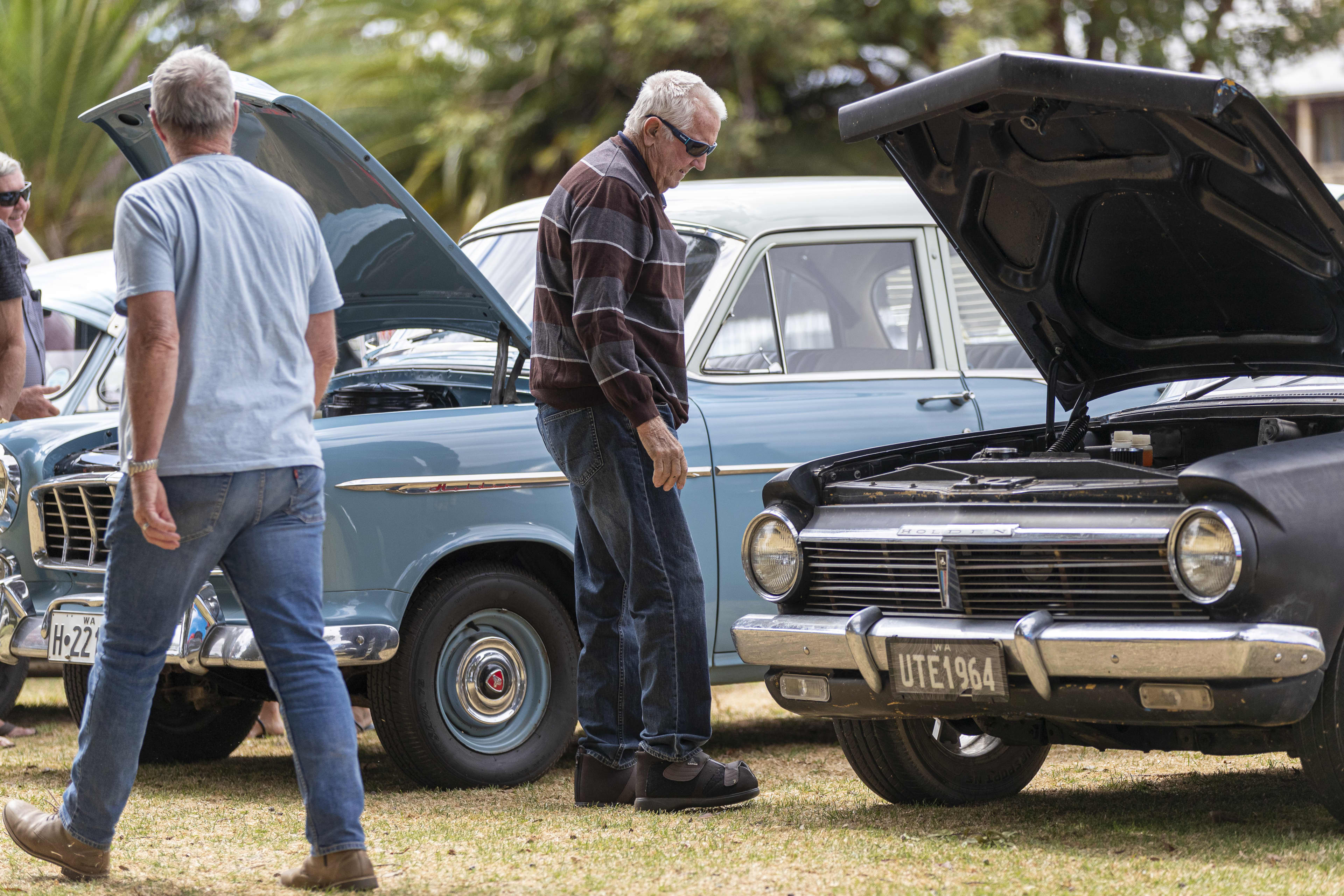 An older man wearing glasses stands beside a light blue vintage car parked outdoors, looking down at the engine with the bonnet open.