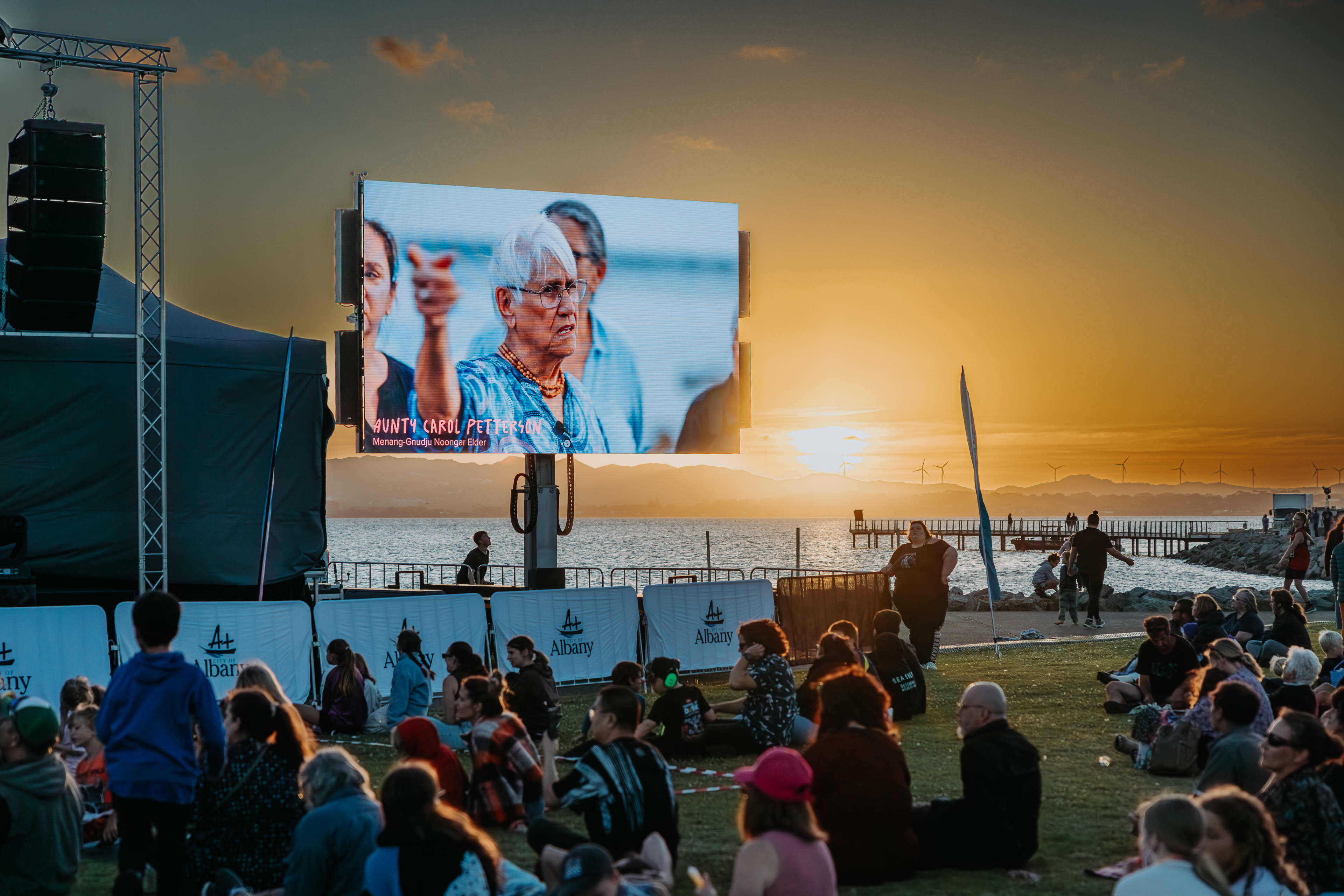 Crowd of people on foreshore with video screen showing stories and sun setting