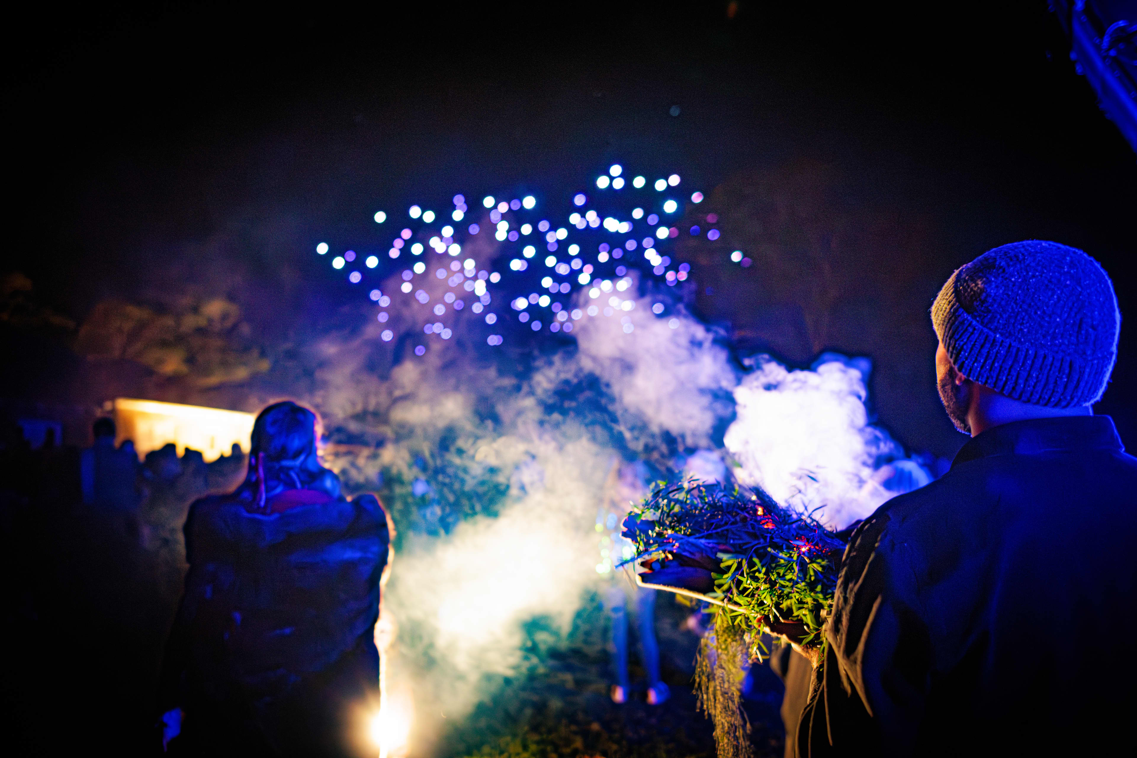 Drone light show with man wearing beanie in foreground carrying smoking sticks and leaves