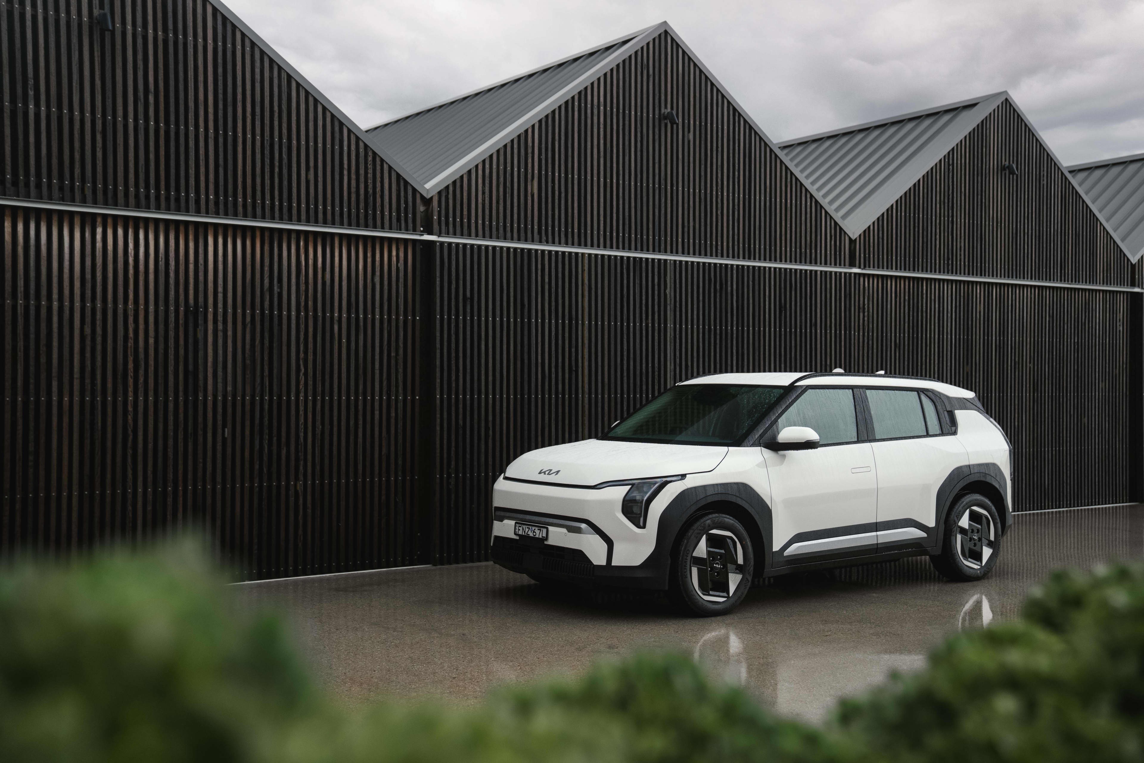 A white Kia Ev3 parked outside wooden sheds
