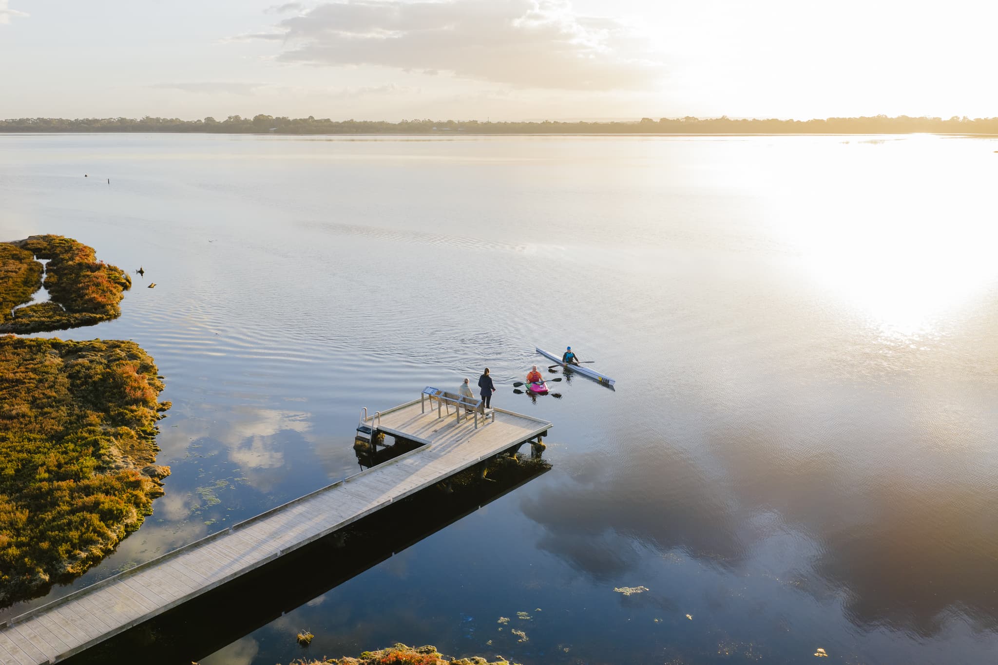An aerial view of two people canoeing at sunrise near a wooden boardwalk, while two others stand on the boardwalk overlooking a calm estuary.