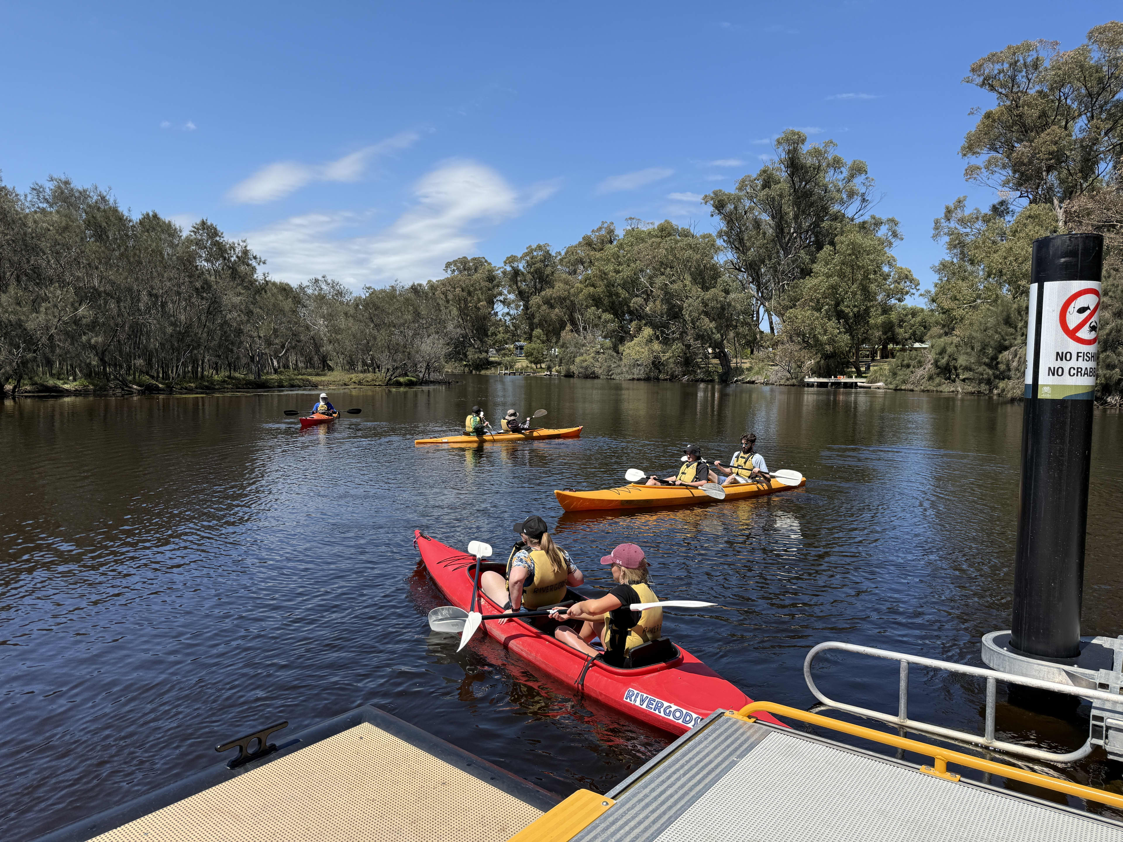 Four canoes with paddlers setting off from a dock into a calm estuary.