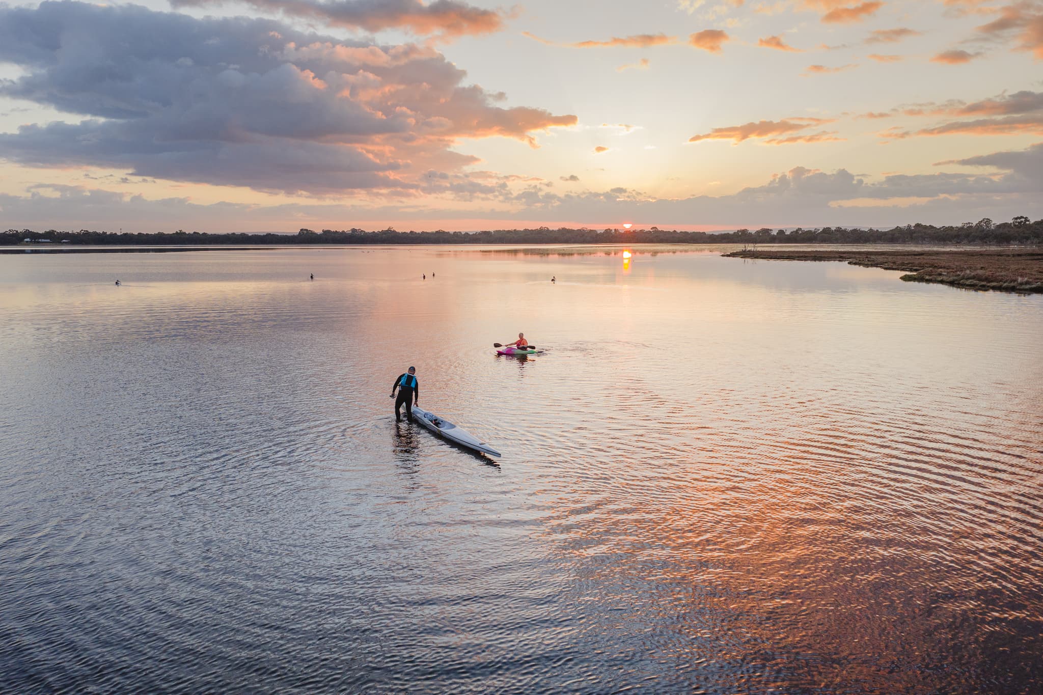 A landscape view of a calm estuary with two paddlers and the sunrise glowing across the water.