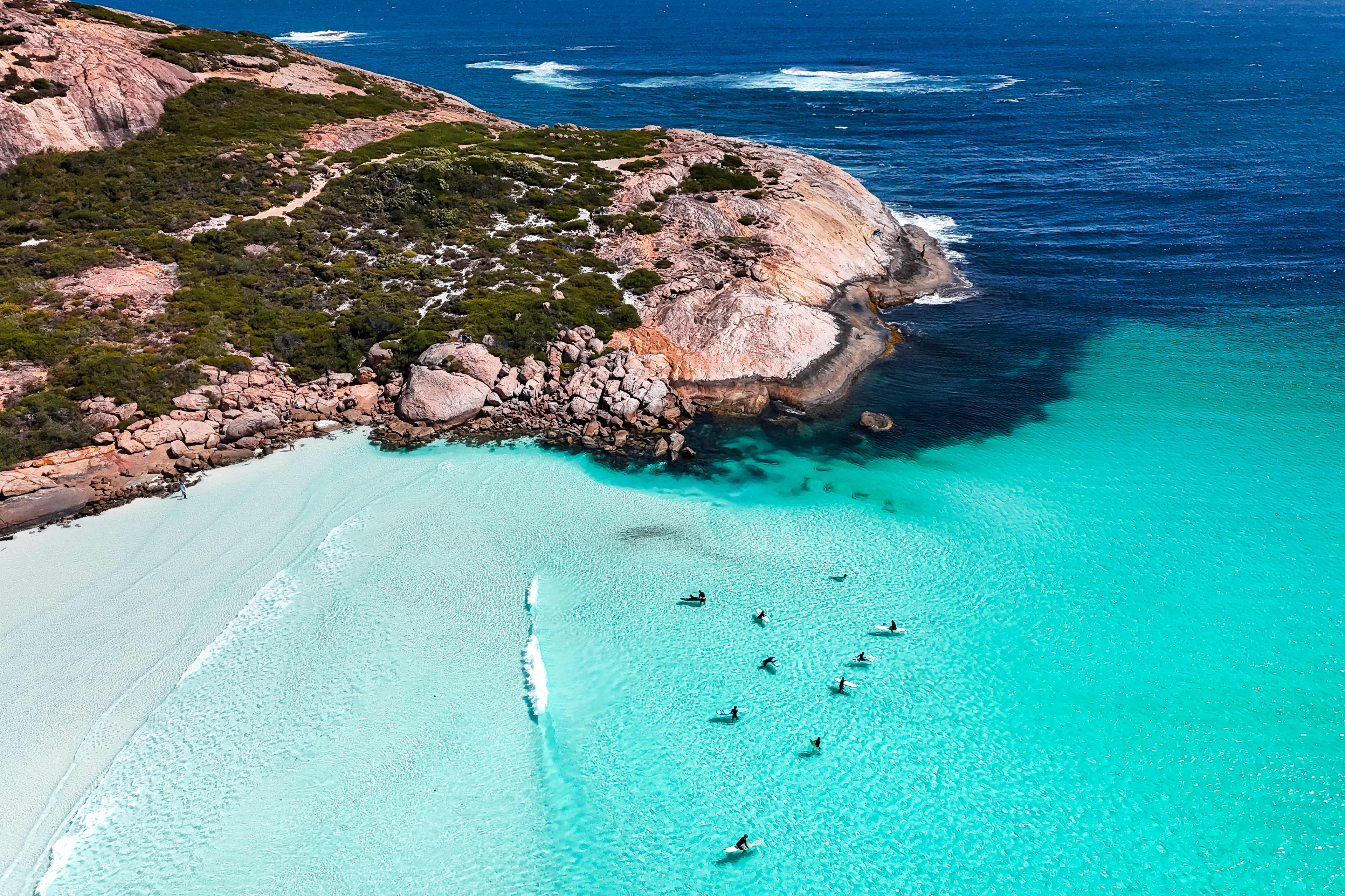 An aerial shot of turquoise blue waters with surfers sitting in the water, with a large granite outcrop creating a protected lagoon.