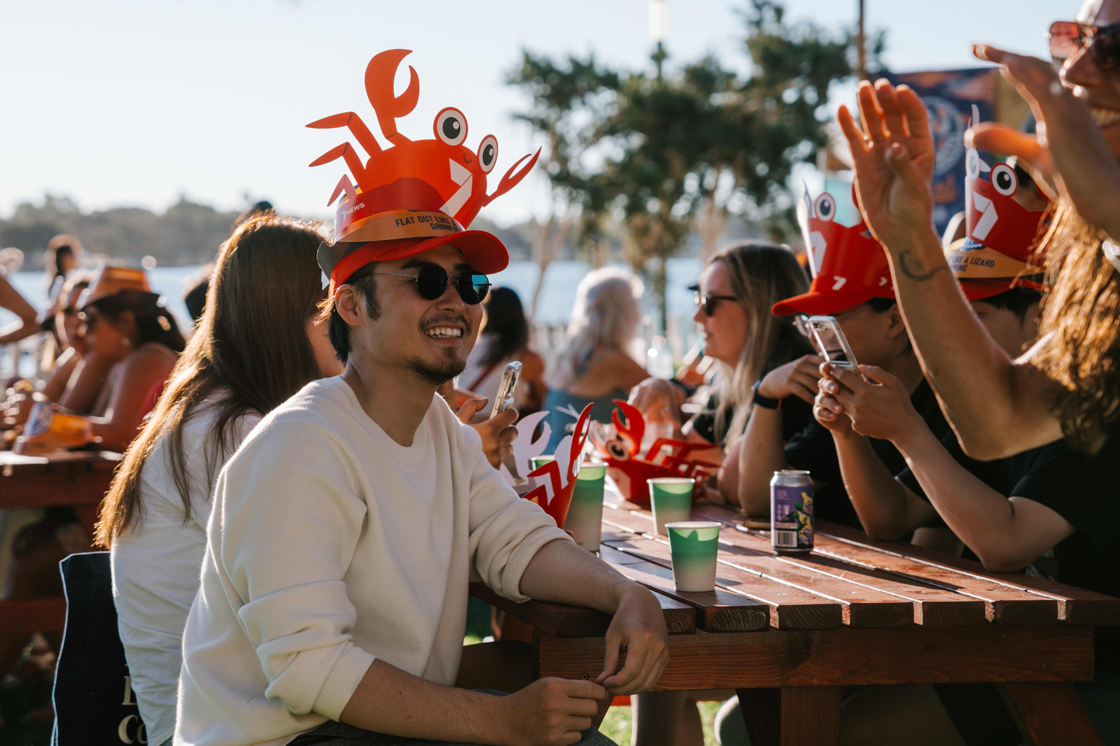 A group of friends sit at an outdoor picnic table by the water on a sunny day, wearing bright red crab-shaped hats and smiling while holding drinks and phones, with trees and the waterfront in the background.