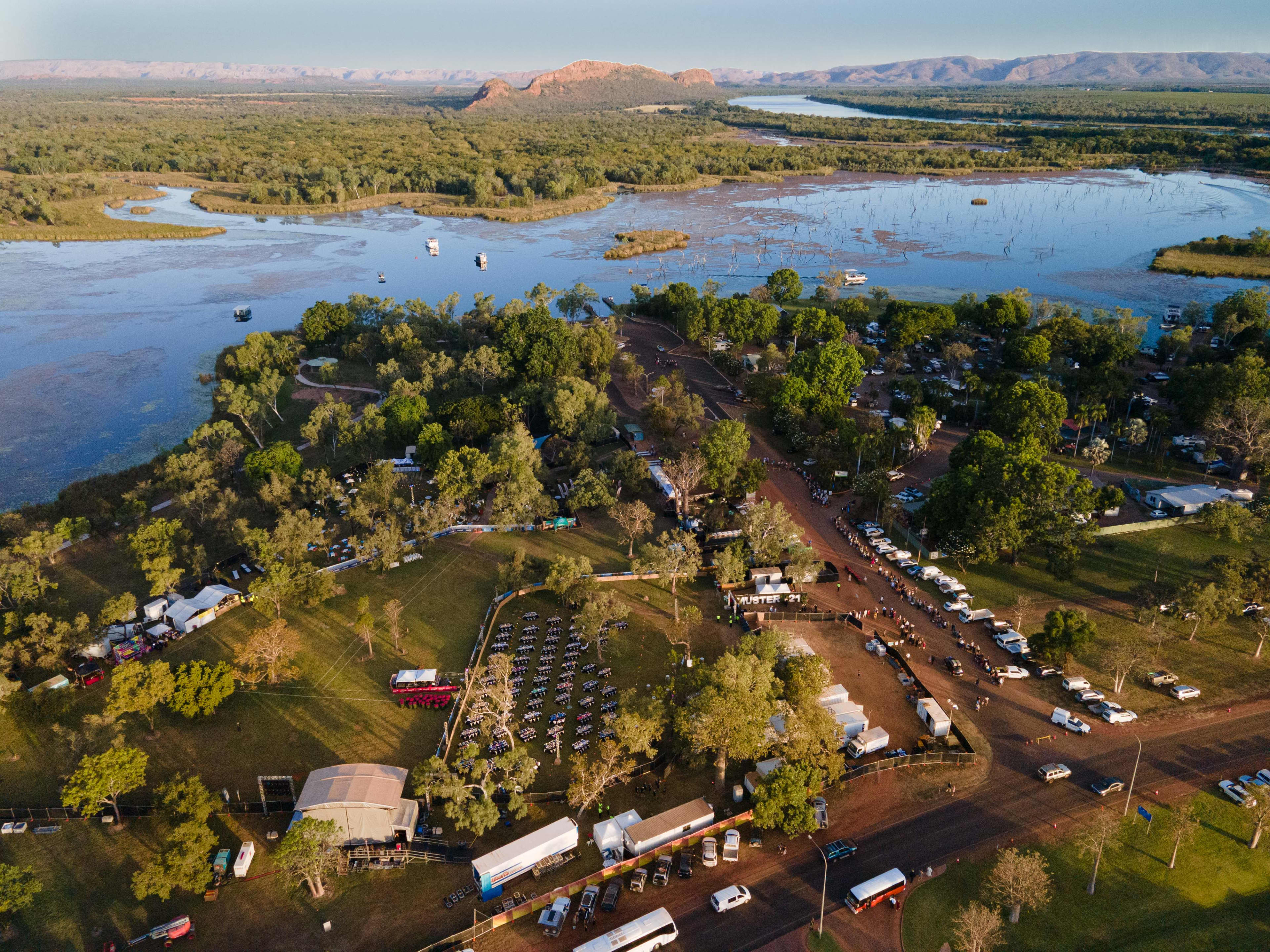 An aerial view of a riverside outdoor event set among trees at sunset, with rows of seating, parked cars, and tents beside a wide waterway surrounded by bushland.