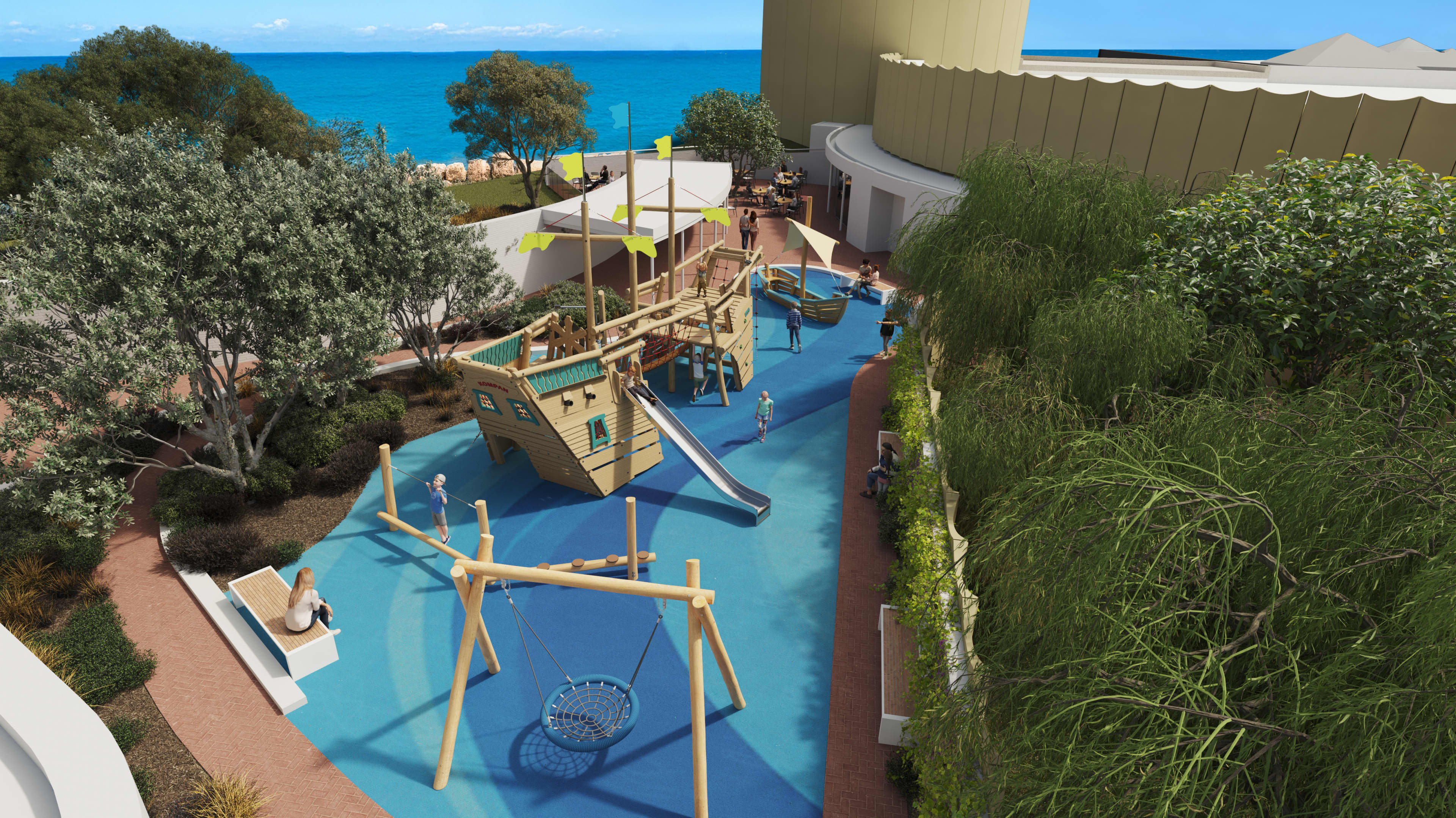 A landscaped seaside playground beside a modern curved building, overlooking bright blue ocean water. The play area features a large wooden pirate ship structure with green flags, climbing nets, and a metal slide, set on blue rubber safety flooring. A basket swing hangs in the foreground, and children play on balance beams and around a small boat feature. Trees and shrubs surround the space, with benches and a shaded seating area where adults sit nearby.