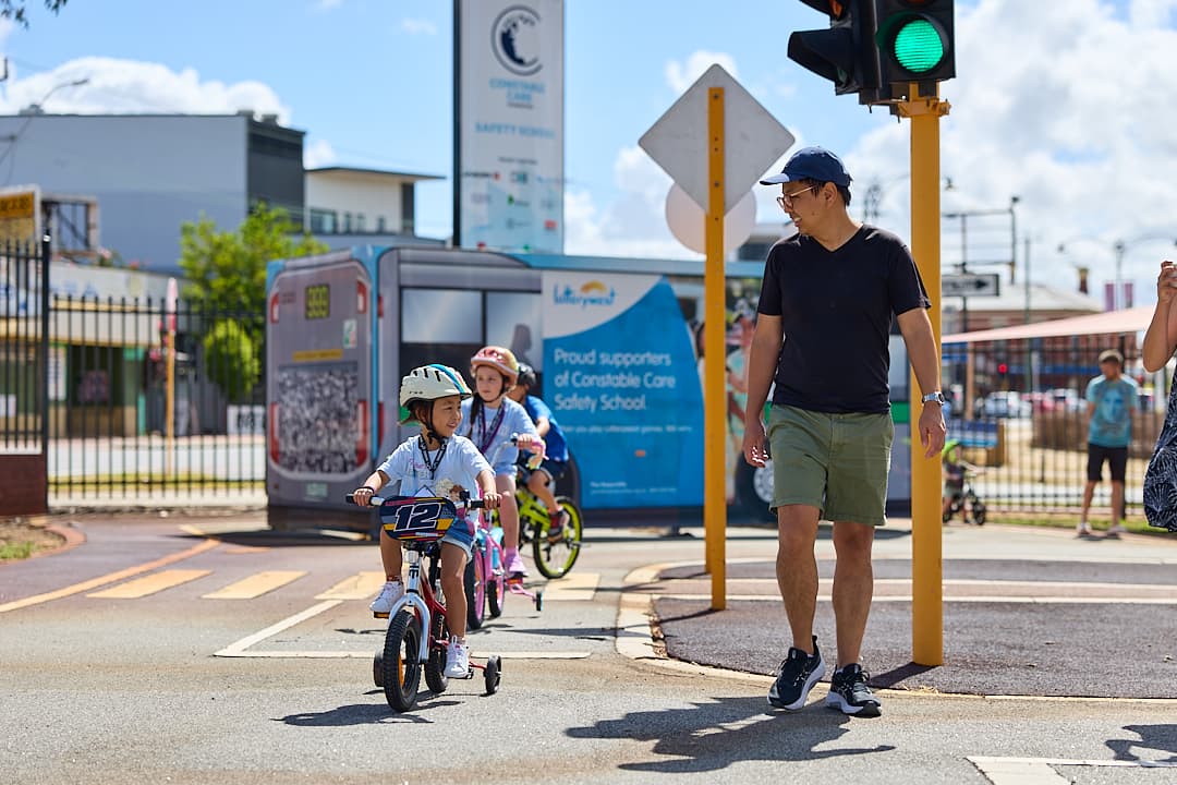 Father walking by his daughter who is on a bike with training wheels and a helmet on her head.