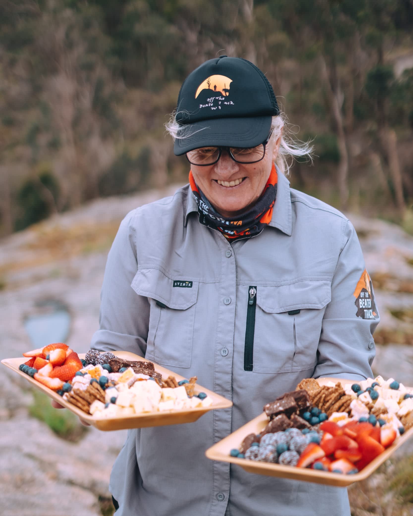 Alt text: A smiling woman wearing glasses, a black cap, and a gray outdoor shirt stands outside on a rocky trail, holding two wooden platters filled with snacks including strawberries, blueberries, cheese cubes, crackers, and chocolate treats.