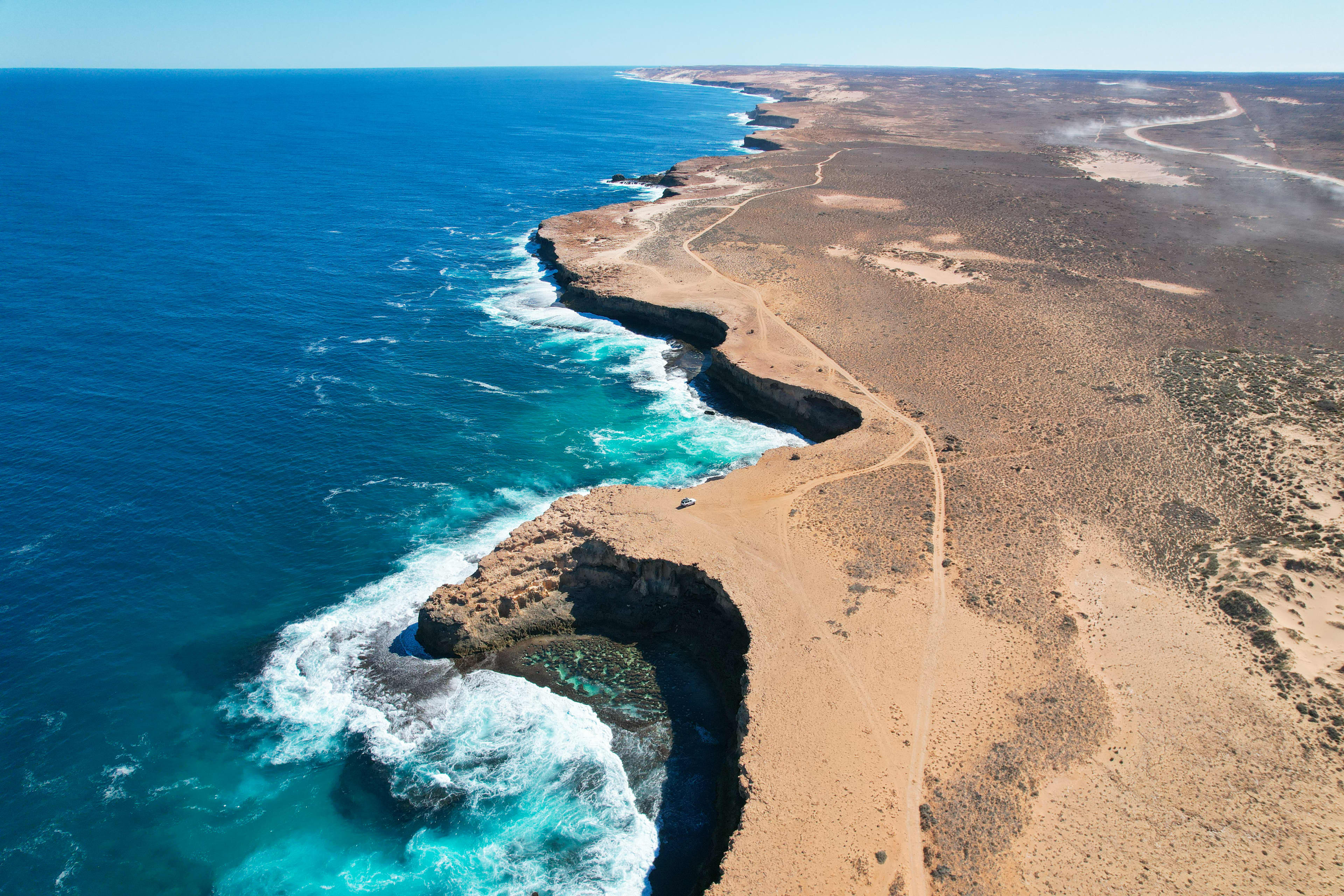 Aerial view of a rugged desert coastline with steep limestone cliffs dropping into bright turquoise ocean waters. Waves crash against the rocky shore, creating white foam, while a dirt track winds along the barren, sandy landscape above the cliffs.