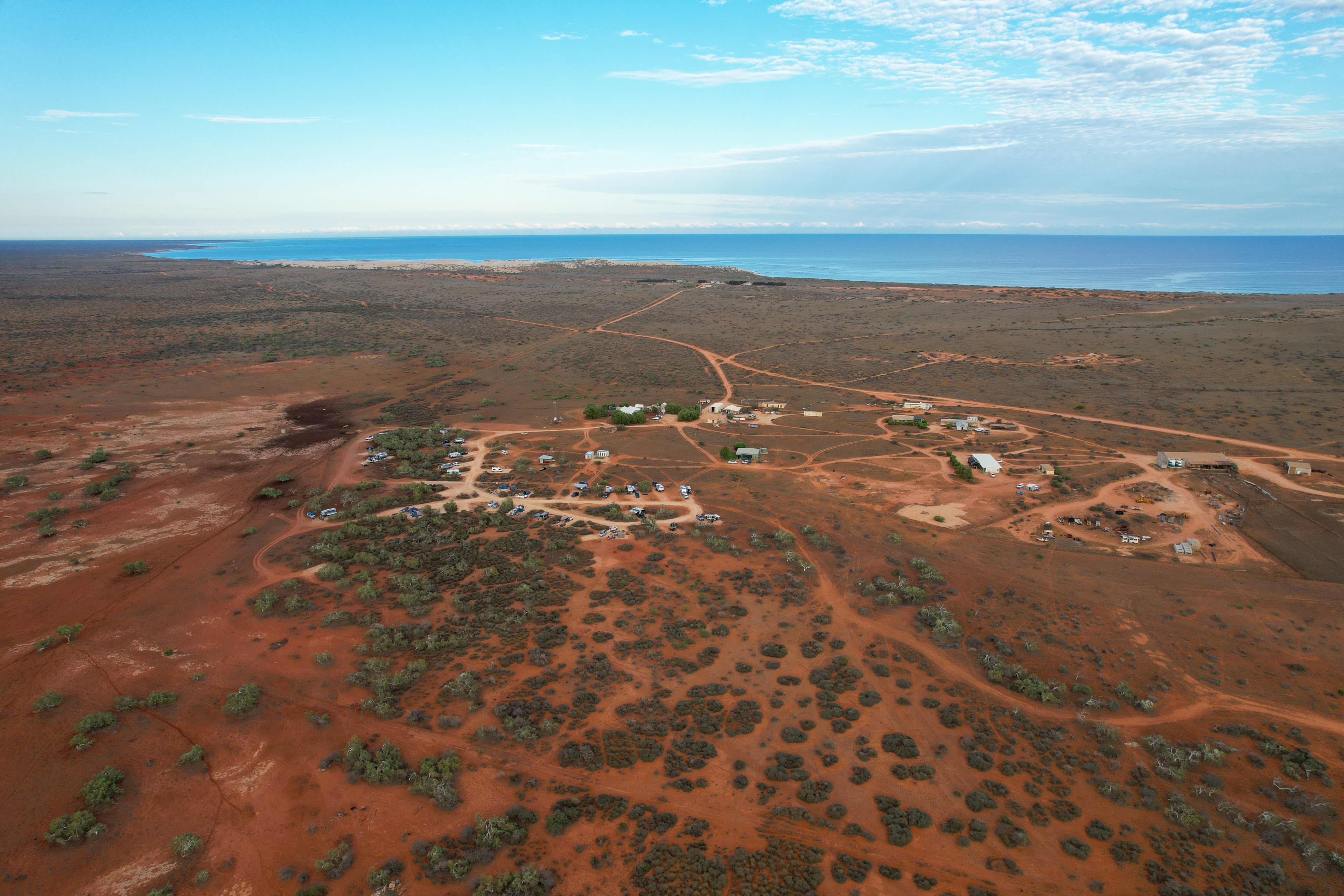 Aerial view of a remote coastal settlement in a red desert landscape, with scattered buildings, caravans, and dirt roads crisscrossing the terrain. Low shrubs dot the ground, and the blue ocean stretches across the horizon under a partly cloudy sky.