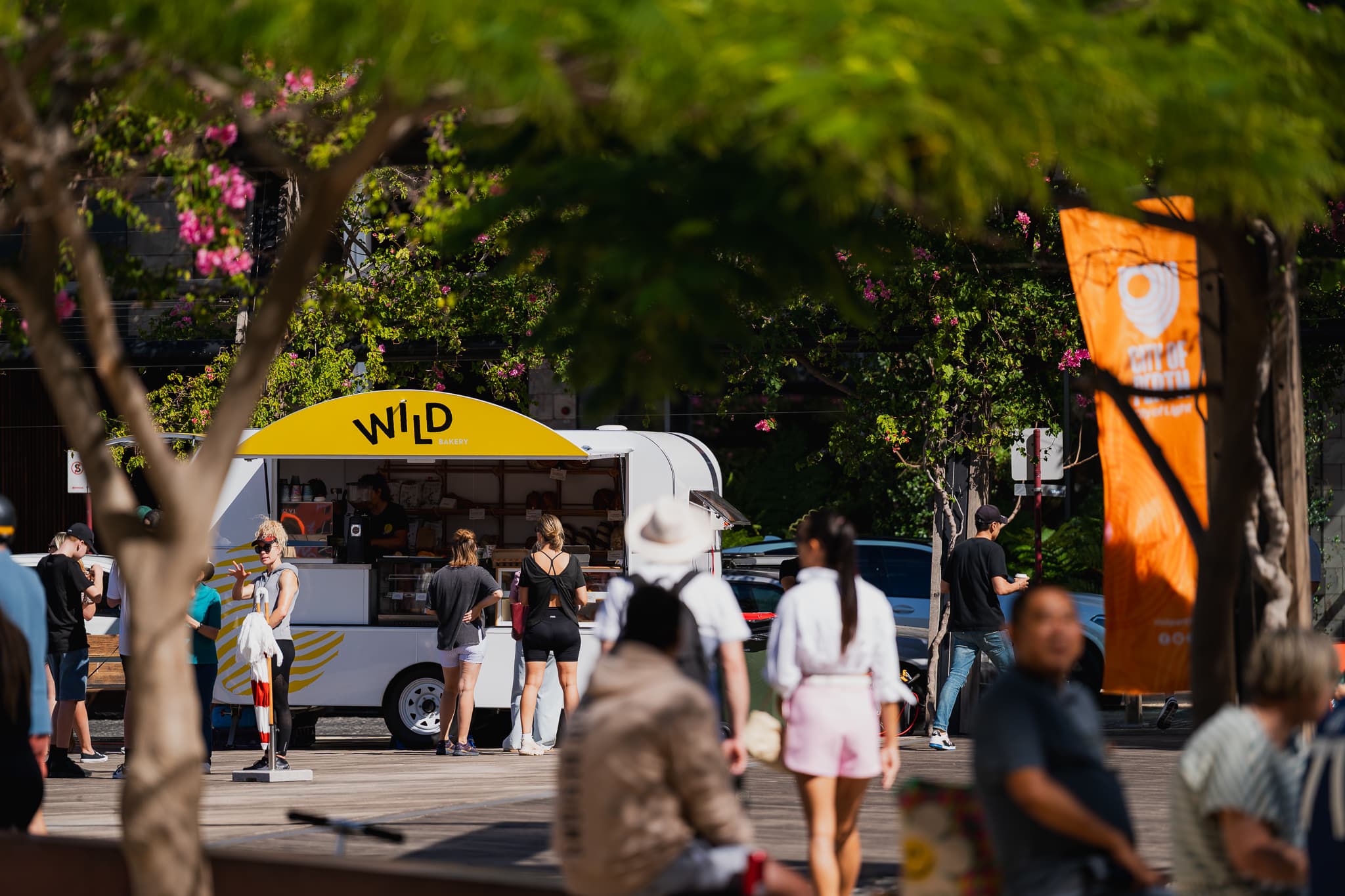 A small white bakery food truck with a bright yellow “WILD” sign serves customers at an outdoor market, surrounded by leafy trees and pink flowers. Several people stand in line while others walk past in the foreground, and an orange vertical banner hangs to the right on a sunny day.