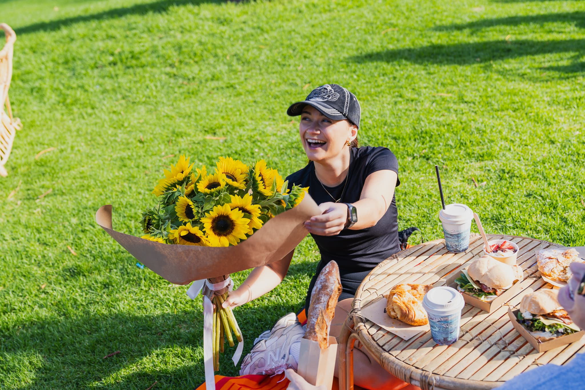 A lady with a black top and cap sitting picnic style in front of a table with breakfast food, holding a bouquet of sunflowers.