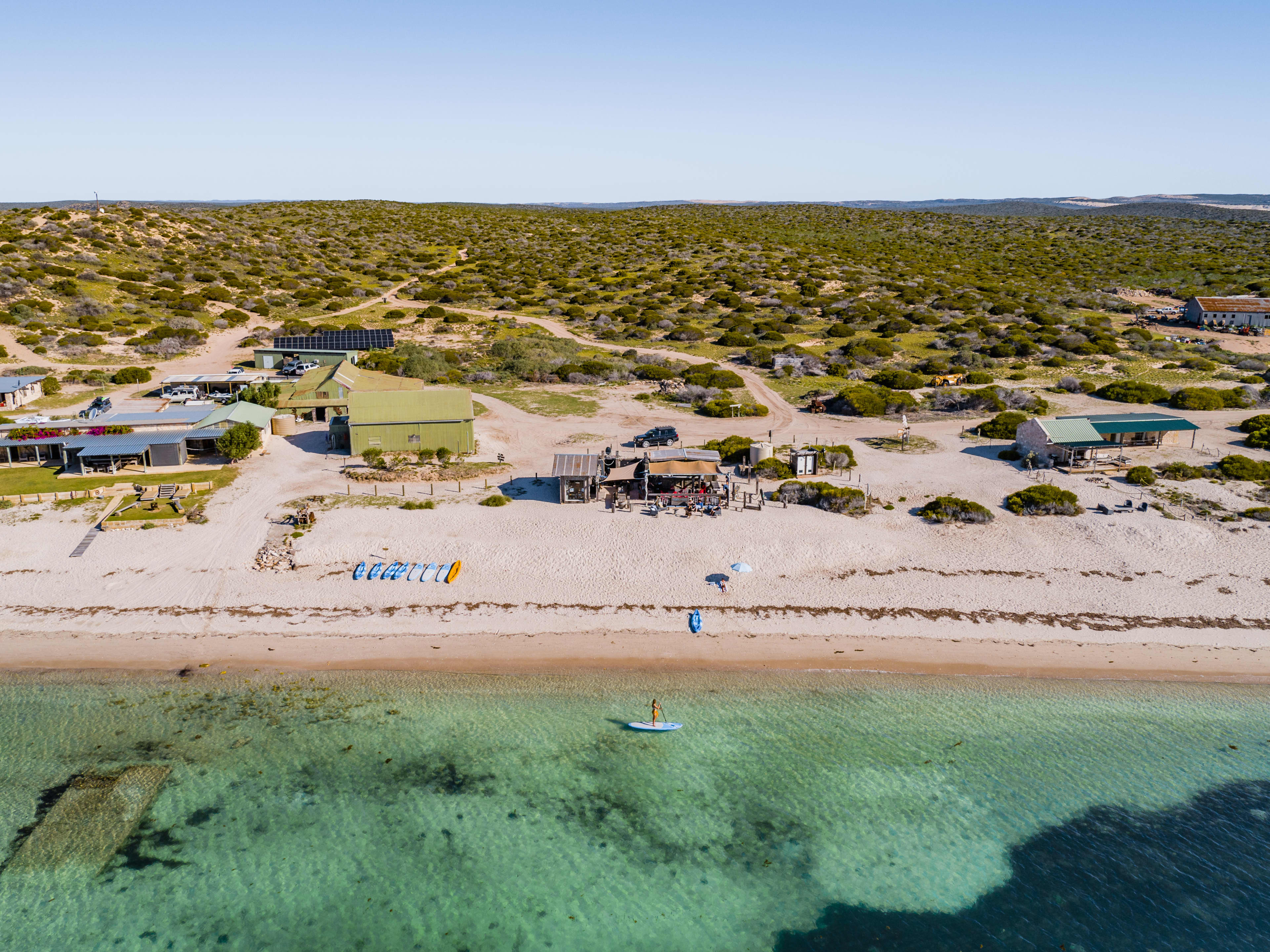 Aerial view of a remote beach with clear turquoise water in the foreground, where a person is stand-up paddleboarding. On the sandy shore, several colorful kayaks are lined up, and a small beachfront café with green-roofed buildings sits just behind the beach. Dirt roads wind through low scrubby vegetation stretching across a flat, arid landscape under a clear blue sky.