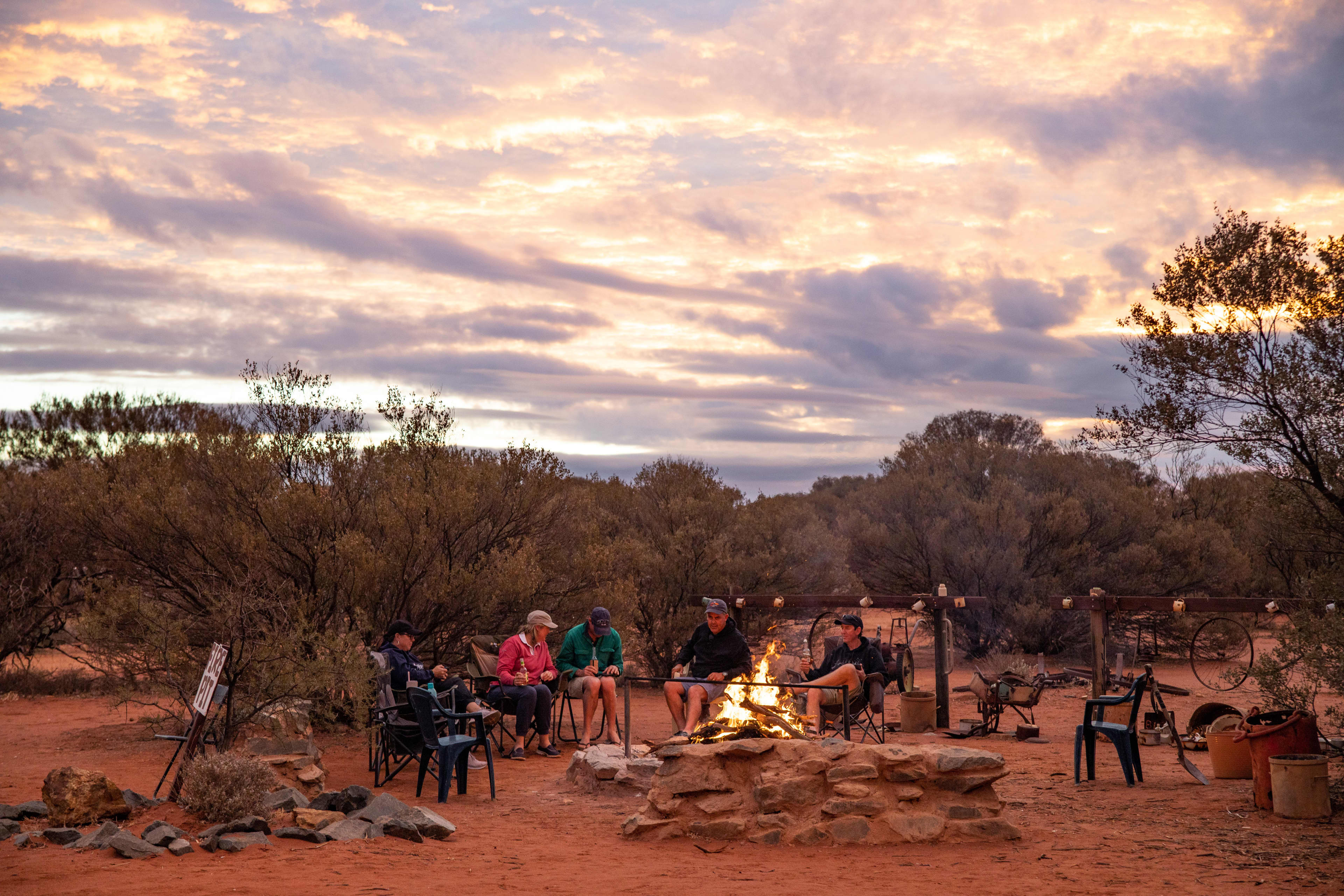 Alt text: Five people sit in camping chairs around a stone fire pit with a small campfire burning at sunset. They are gathered in a red dirt outback setting surrounded by low trees and scrub, with old station equipment and buckets nearby. The sky is filled with pink and orange clouds as evening light fades over the bush landscape.