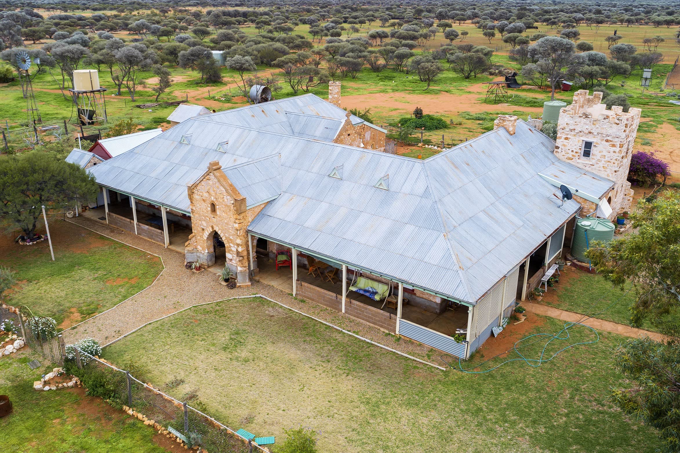Aerial view of a large stone farmhouse with a corrugated metal roof and wraparound veranda, surrounded by fenced lawn and outbuildings. Water tanks and windmills stand nearby, and the property is set within a dry, sparsely wooded rural landscape with scattered trees stretching into the distance.