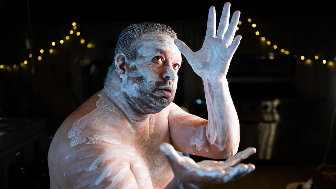 A shirtless man covered in white body paint poses dramatically with one arm raised and the other extended across his chest, under warm string lights at night.
