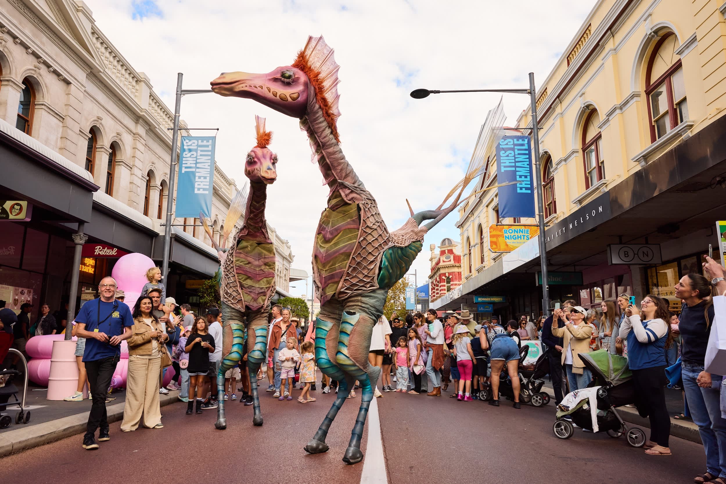 Two tall stilt performers dressed in colourful, dinosaur-like costumes walk down a busy city street during a festival.