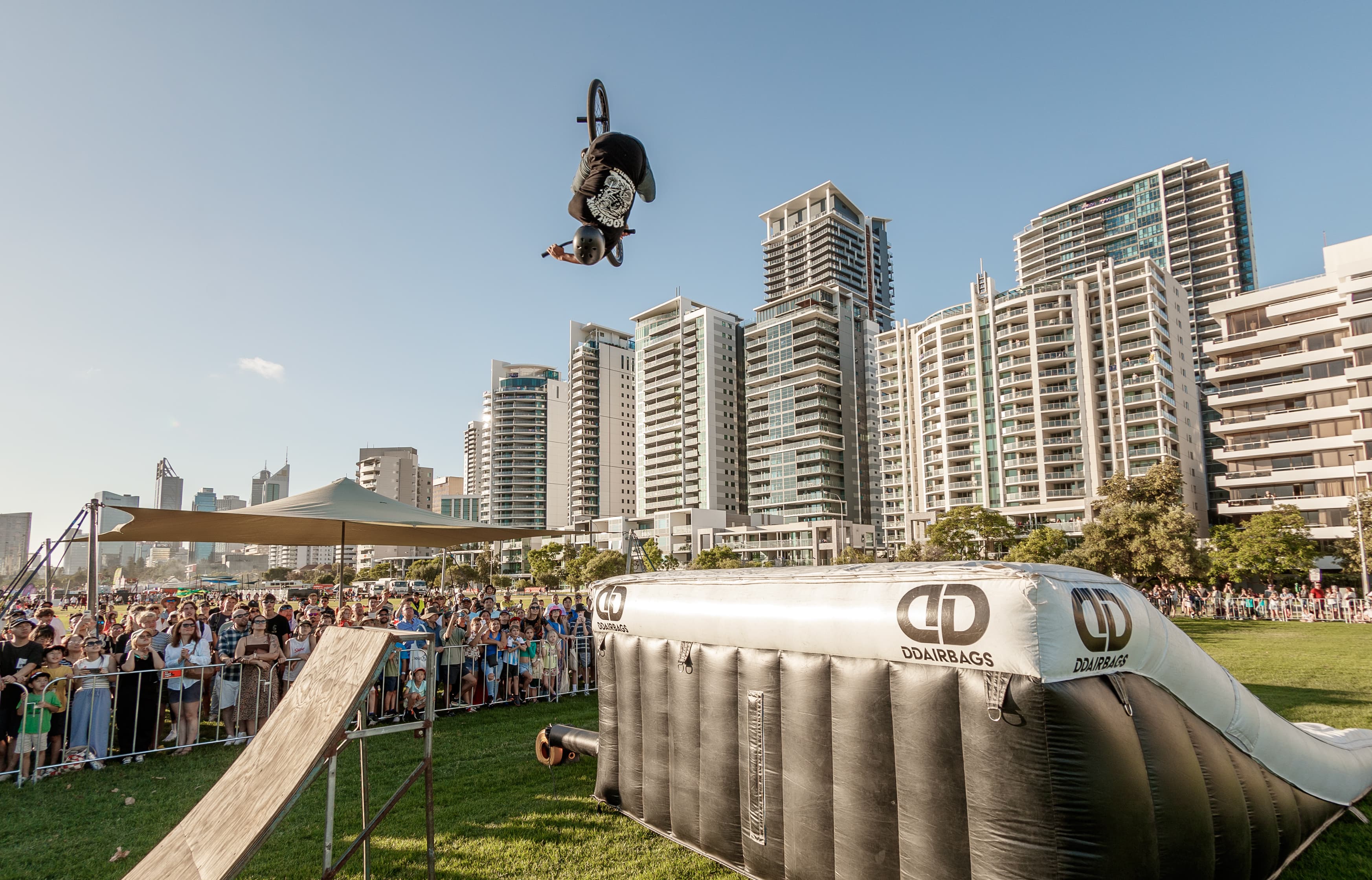 A BMX rider wearing a helmet performs a high backflip above a large inflatable landing pad while a crowd watches from behind barriers in a grassy urban park, with tall apartment buildings and a clear blue sky in the background.