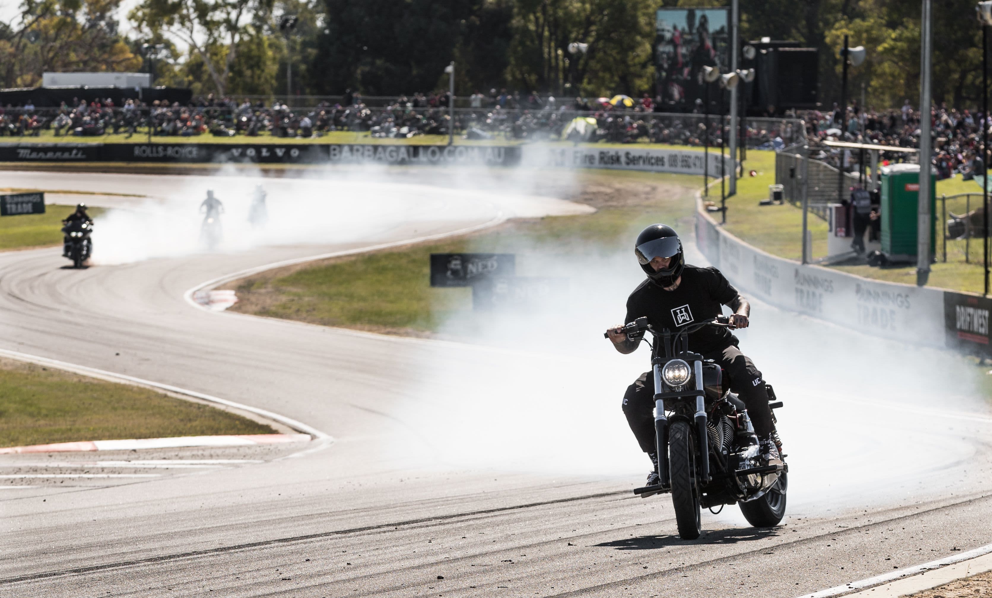A motorcyclist wearing a black helmet rides along a racetrack while performing a smoky burnout, with other riders in the distance and a large crowd of spectators watching from behind barriers.