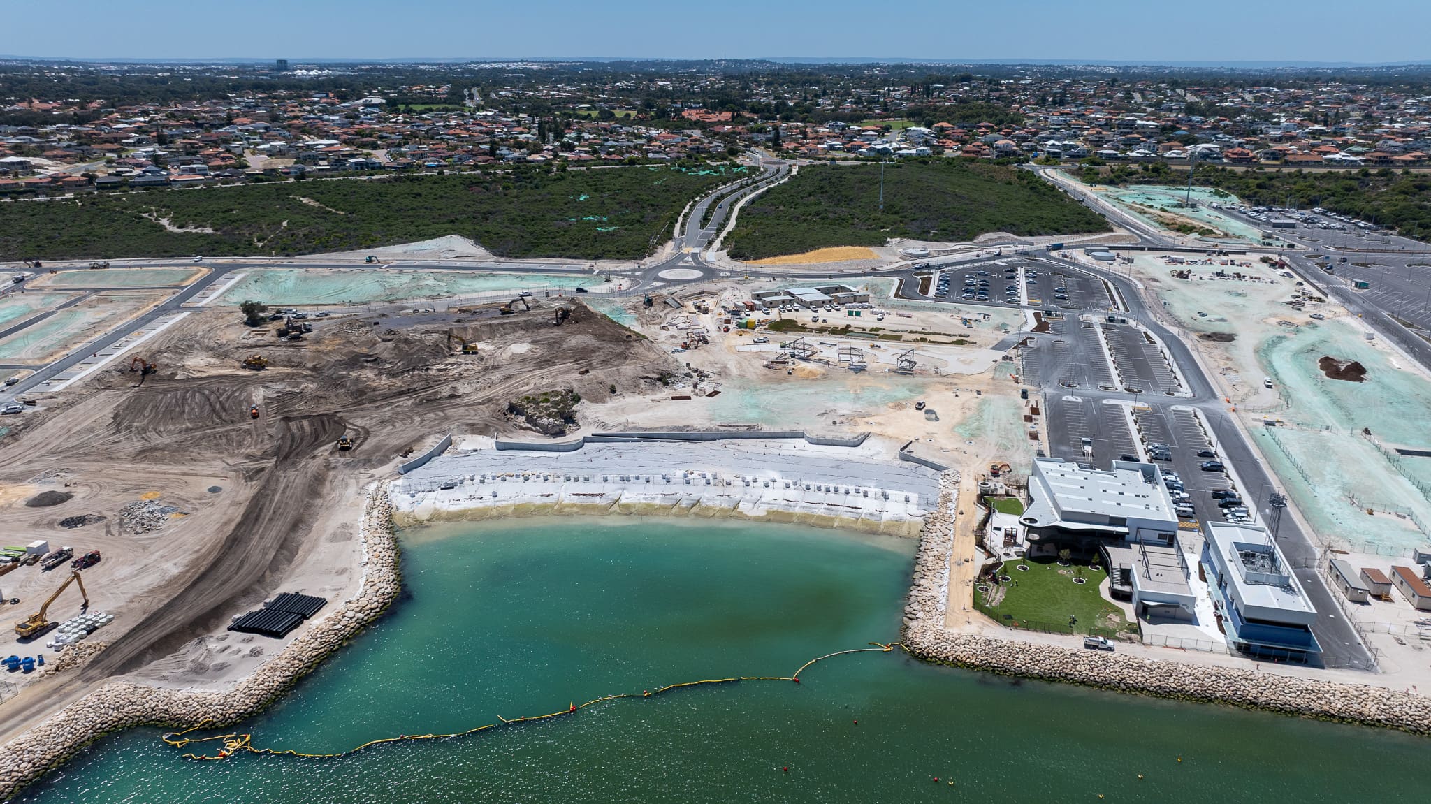 Aerial view of a coastal construction site near a populated area; land grading in progress, adjacent to a calm waterfront with blue-green sea.