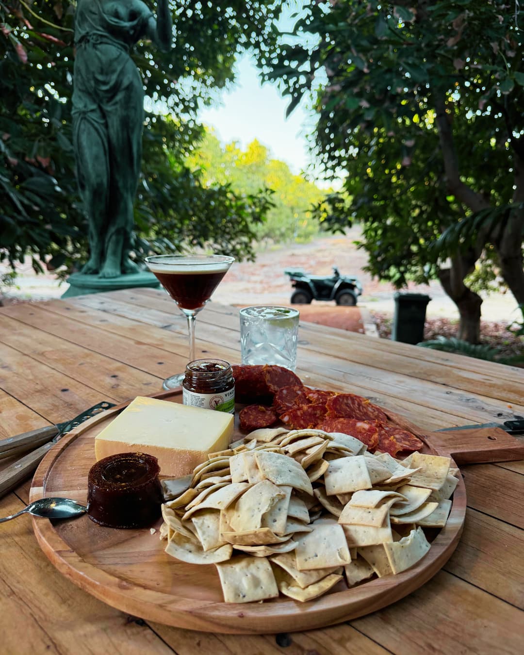 Food platter accompanied by glasses of spirits on shady bench beneath trees.