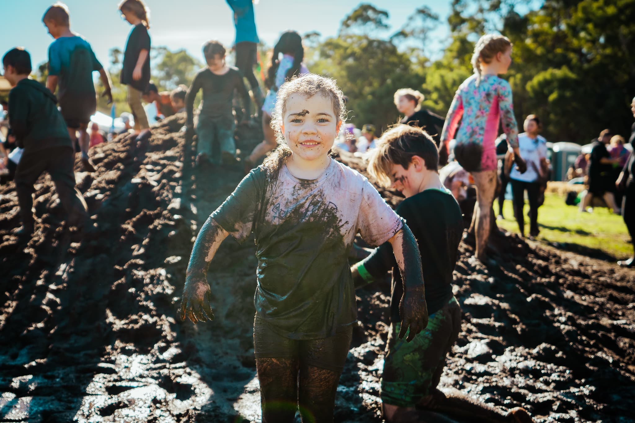 Girl covered in mud at outdoors event with other muddy children in background.