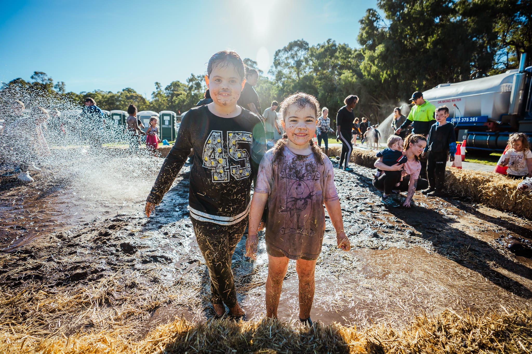 Two muddy children in mud pit with grass and other muddy children in background.