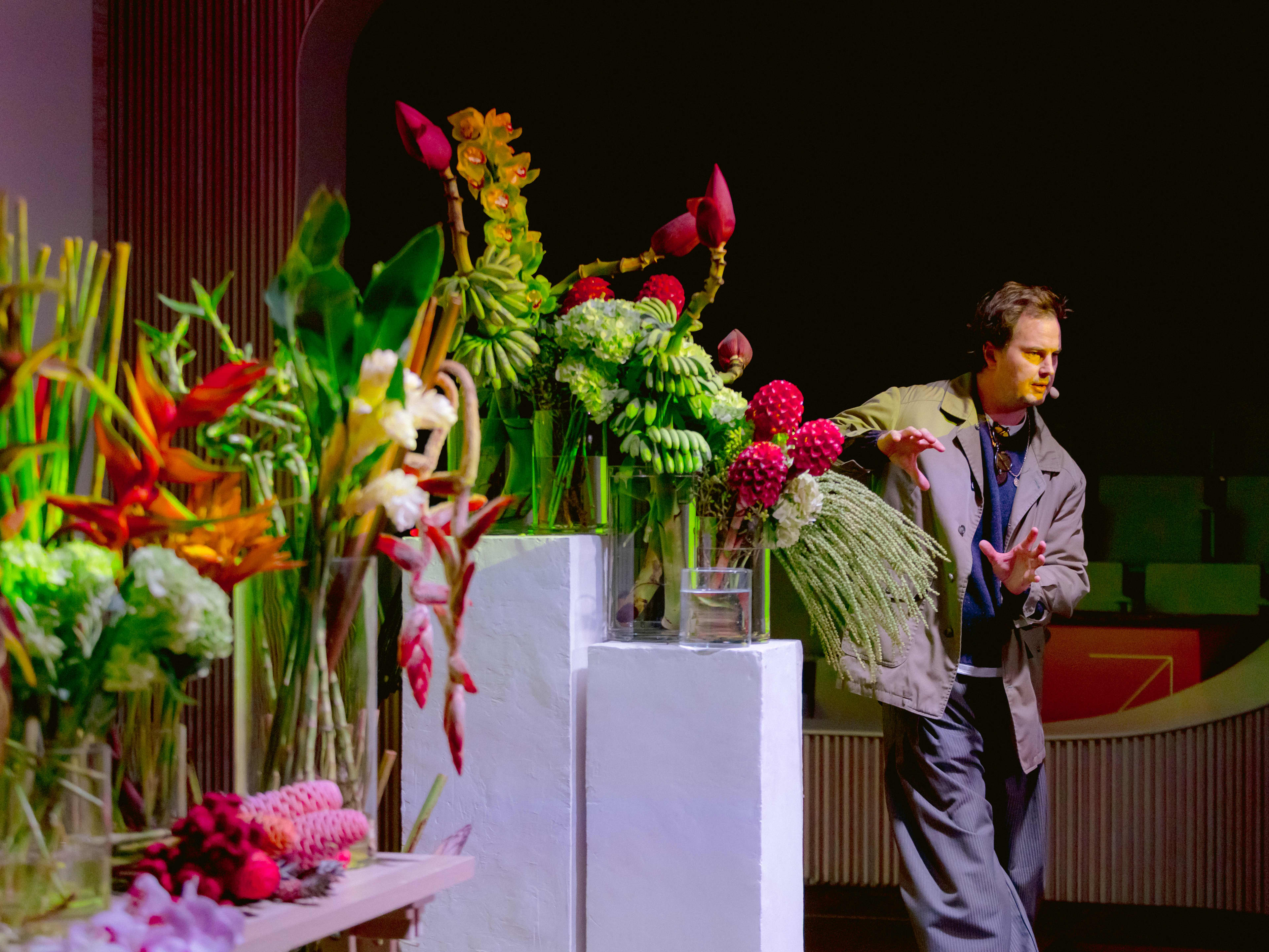 Man standing in front of table full of floral arrangements with microphone.