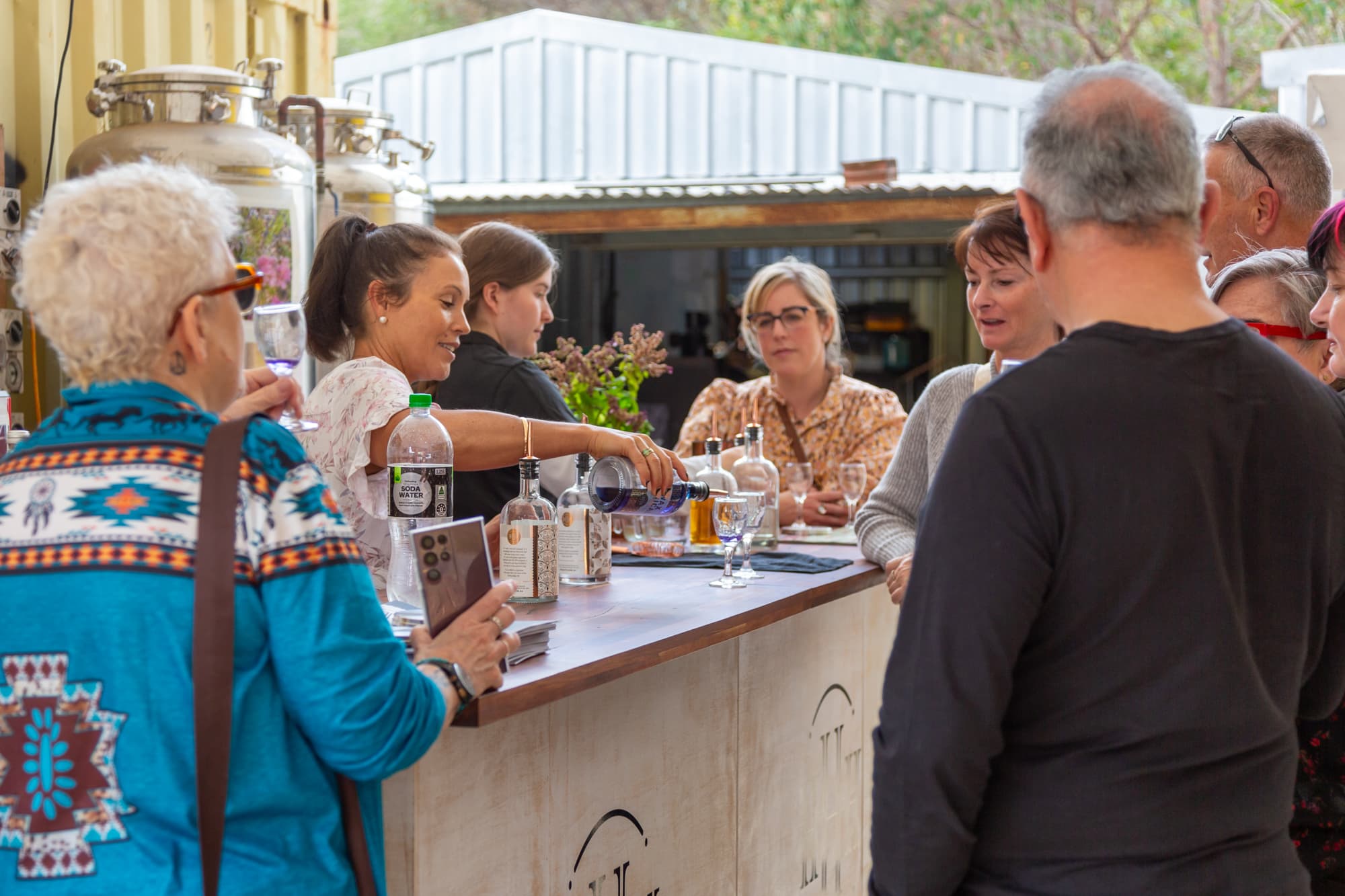 A group of people standing around a wooden tasting station, with a brunette lady pouring a purple spirit into a tasting glass.