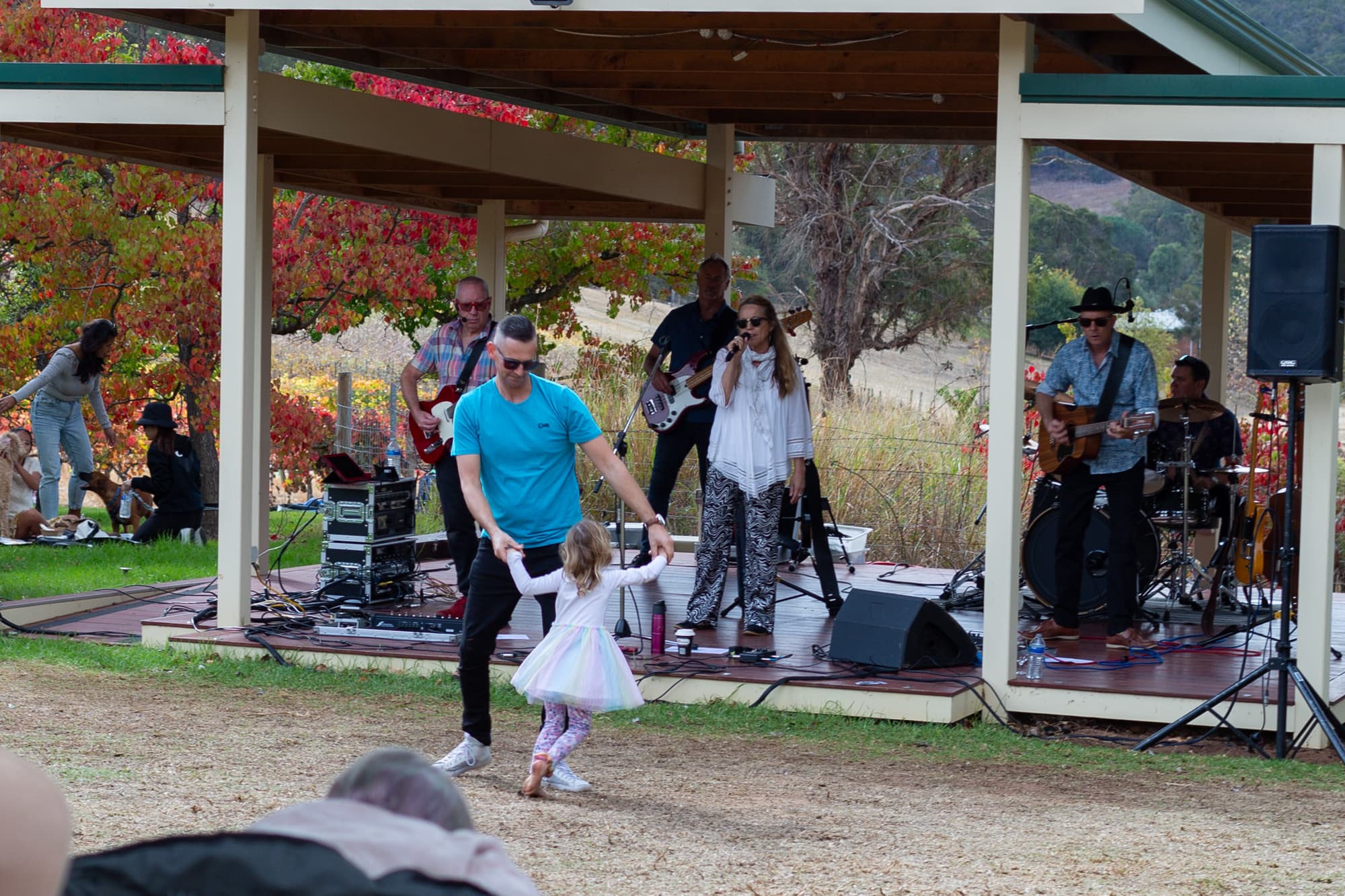 A women singing with 4 men playing guitars and drums behind her at an outdoor venue, with a man and a young child dancing.