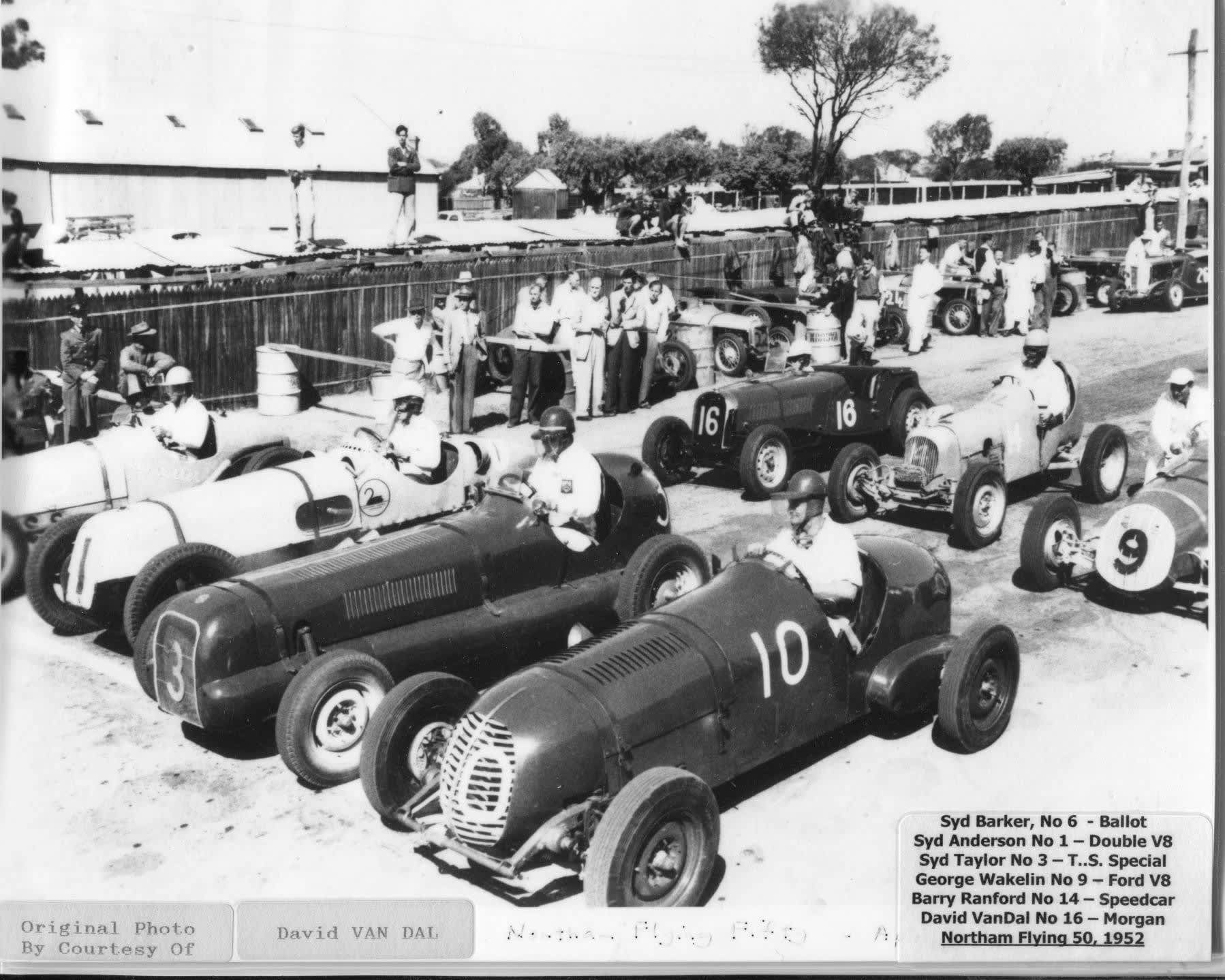Black-and-white historical photograph of a lineup of vintage open-wheel race cars parked side by side in a pit or staging area. Drivers wearing helmets sit in several of the cars, while a group of spectators and crew members stand behind a wooden fence watching. The cars have large painted numbers on their sides (including 3, 10, and 16) and exposed wheels typical of early mid-20th-century racing. In the background are low buildings, trees, and a few people perched on the fence or roof for a better view. A caption in the lower corner identifies drivers, car types, and the event as the Northam Flying 50 in 1952.