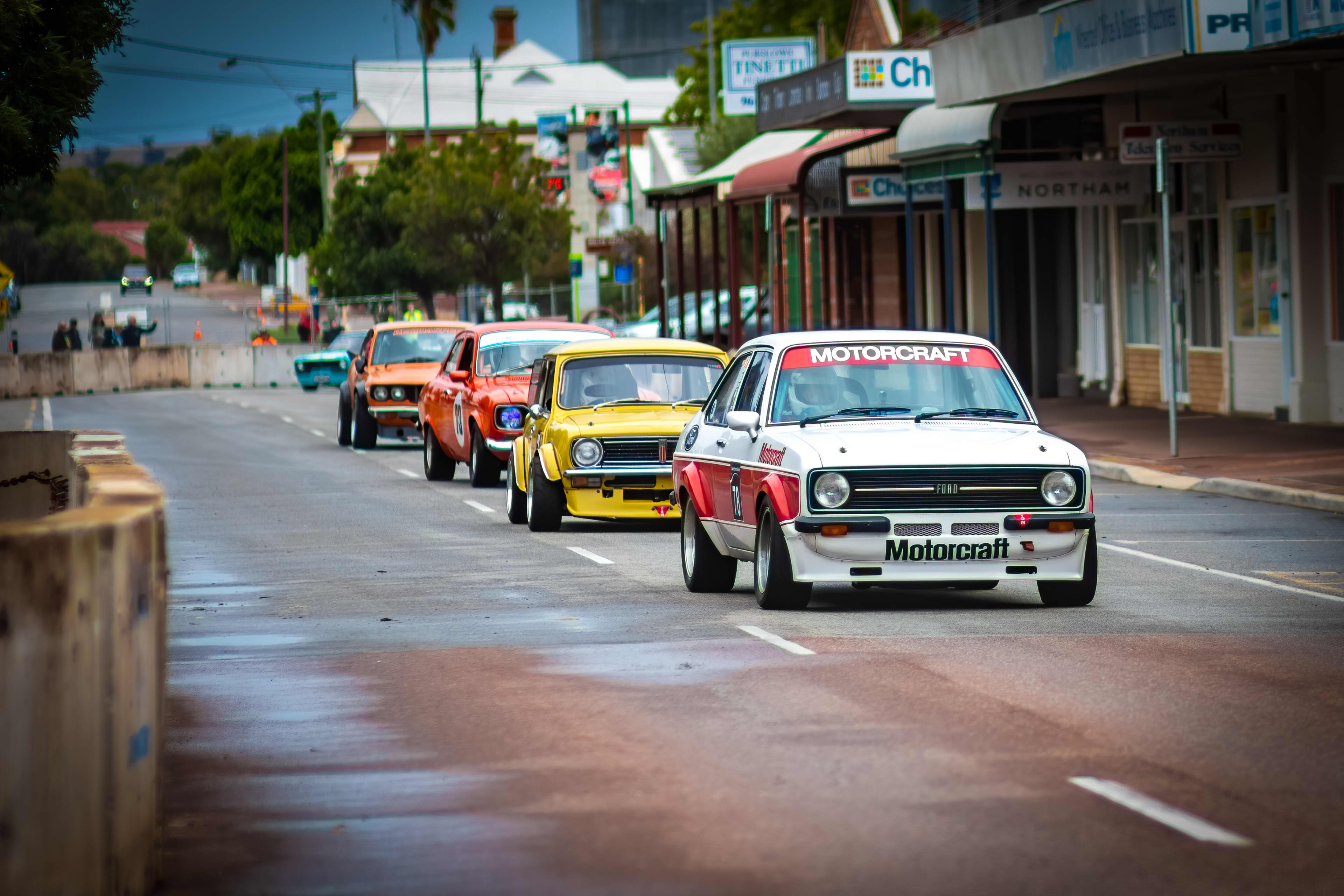 Color photograph of a group of vintage race cars driving in a line down a closed-off town street during a motorsport event. A white Ford sedan with red accents and “Motorcraft” branding leads, followed closely by yellow, orange, and green classic cars.