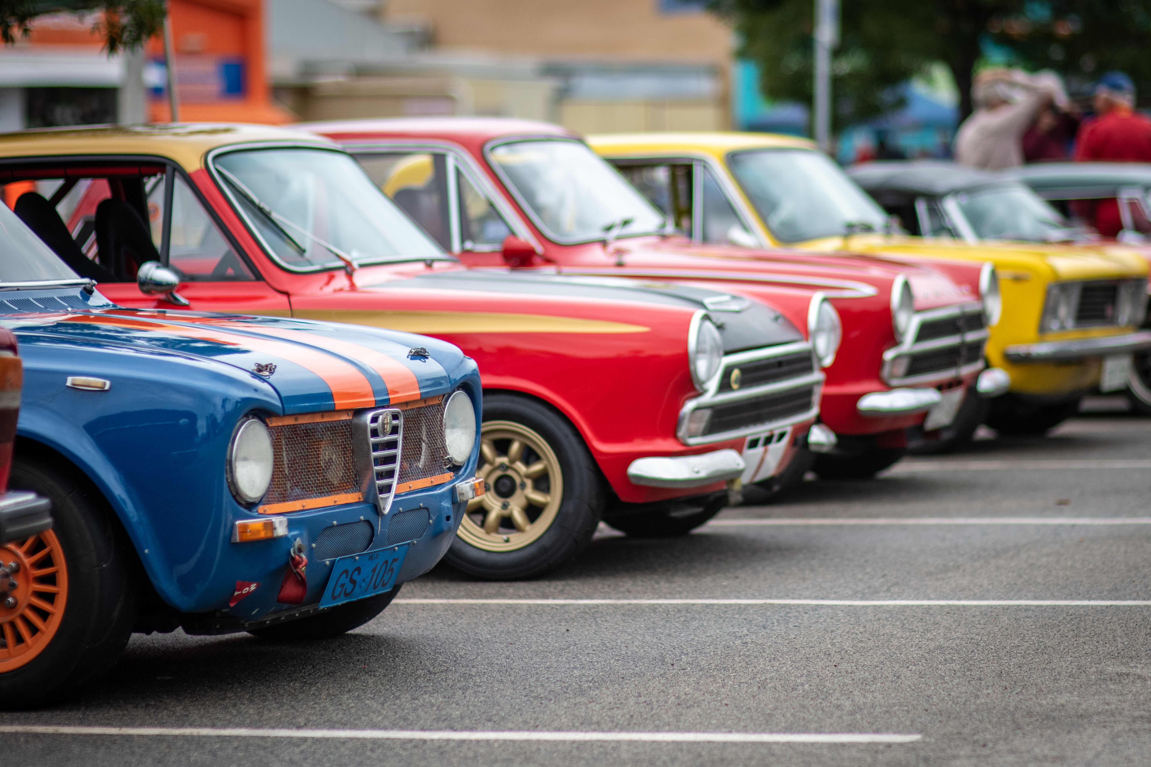 Colour photograph of a row of vintage classic cars parked diagonally in a lot, viewed from the front-left angle. In the foreground, a blue car with orange racing stripes and a distinctive triangular grille stands out, followed by red and yellow cars with chrome details and period styling.