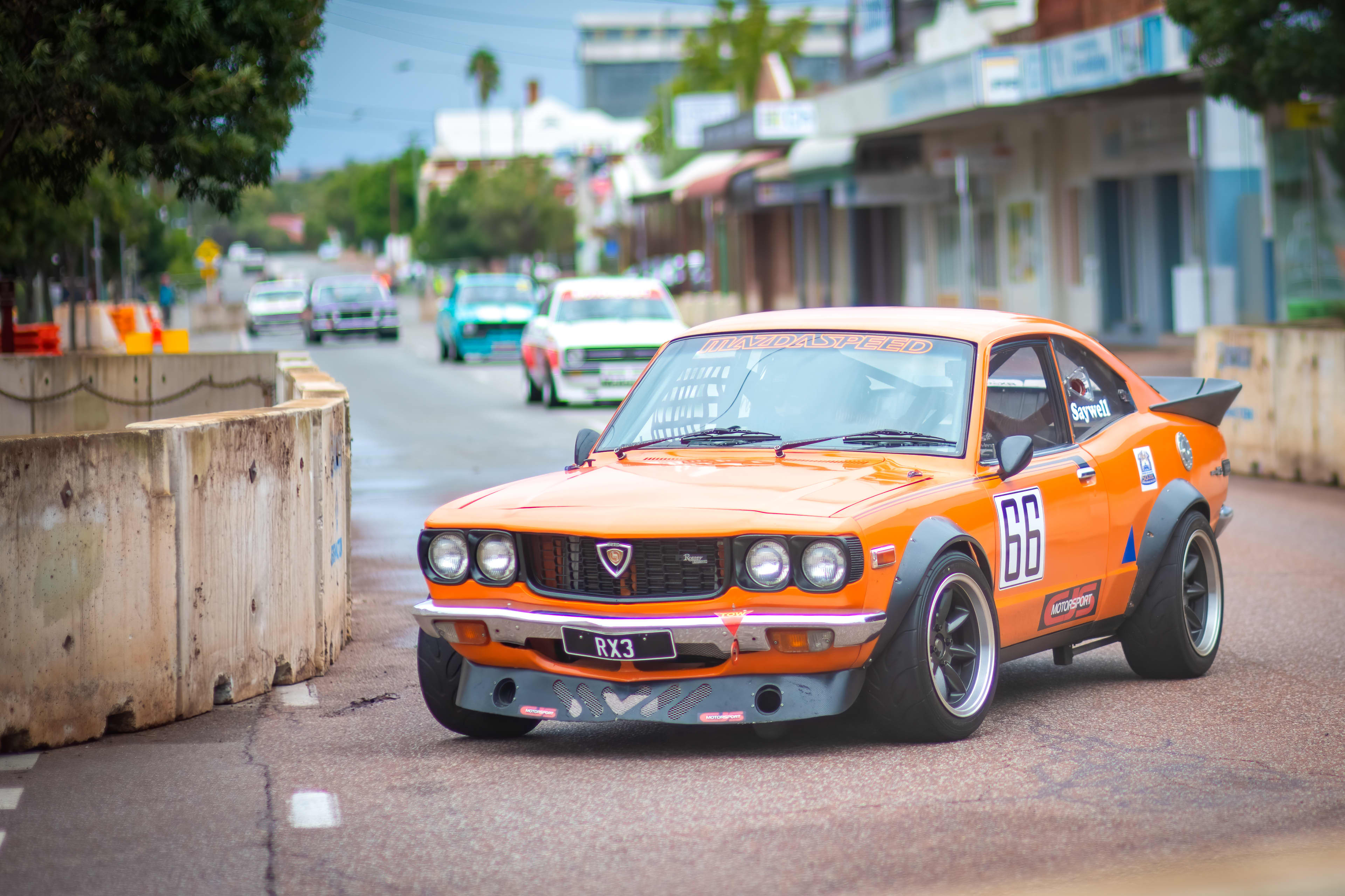 A bright orange race car with 'Mazda Speed' racing down a town streetscape.
