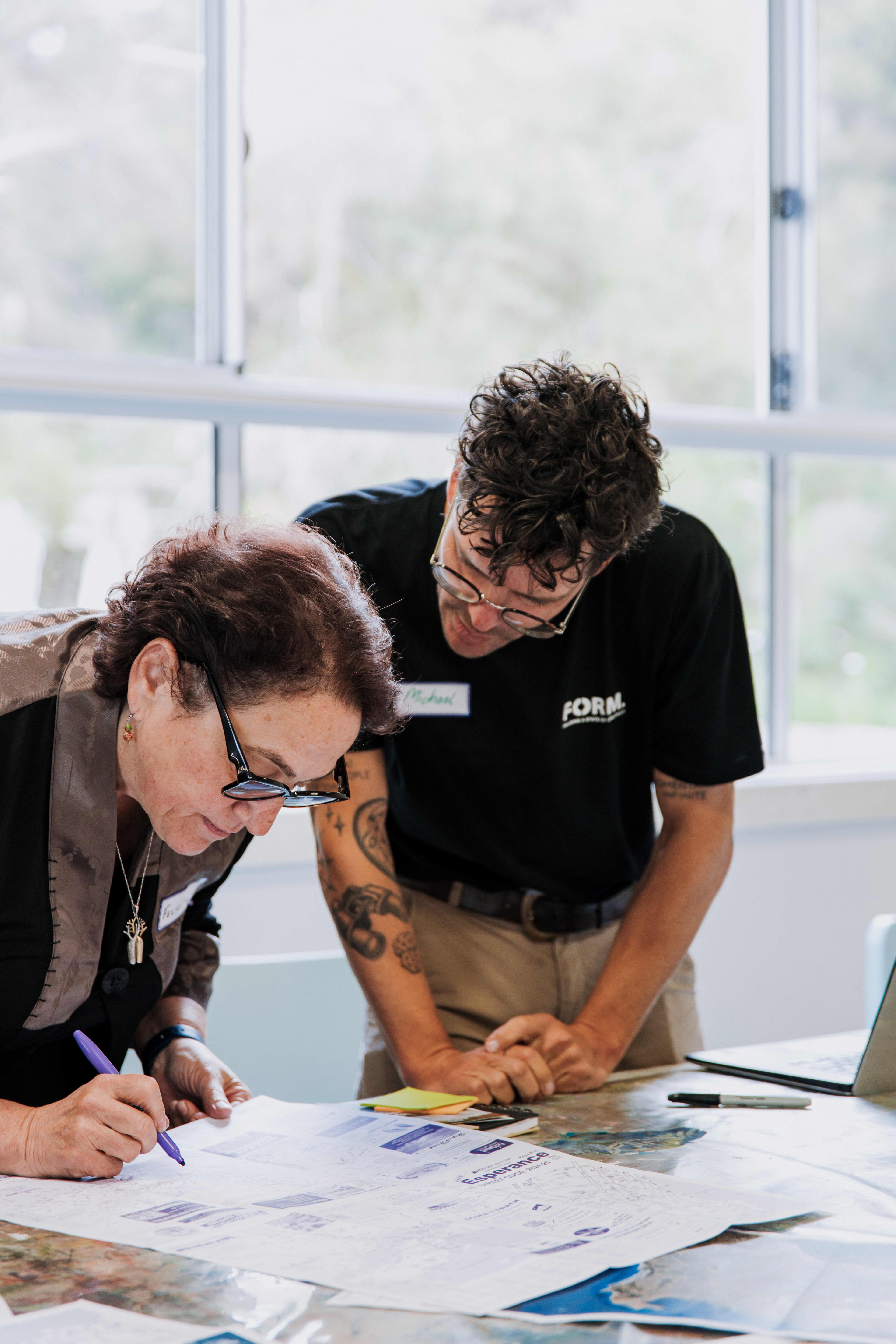 A brunette lady with black glasses marking a map, with a brunette male with a 'FORM' shirt overlooking.