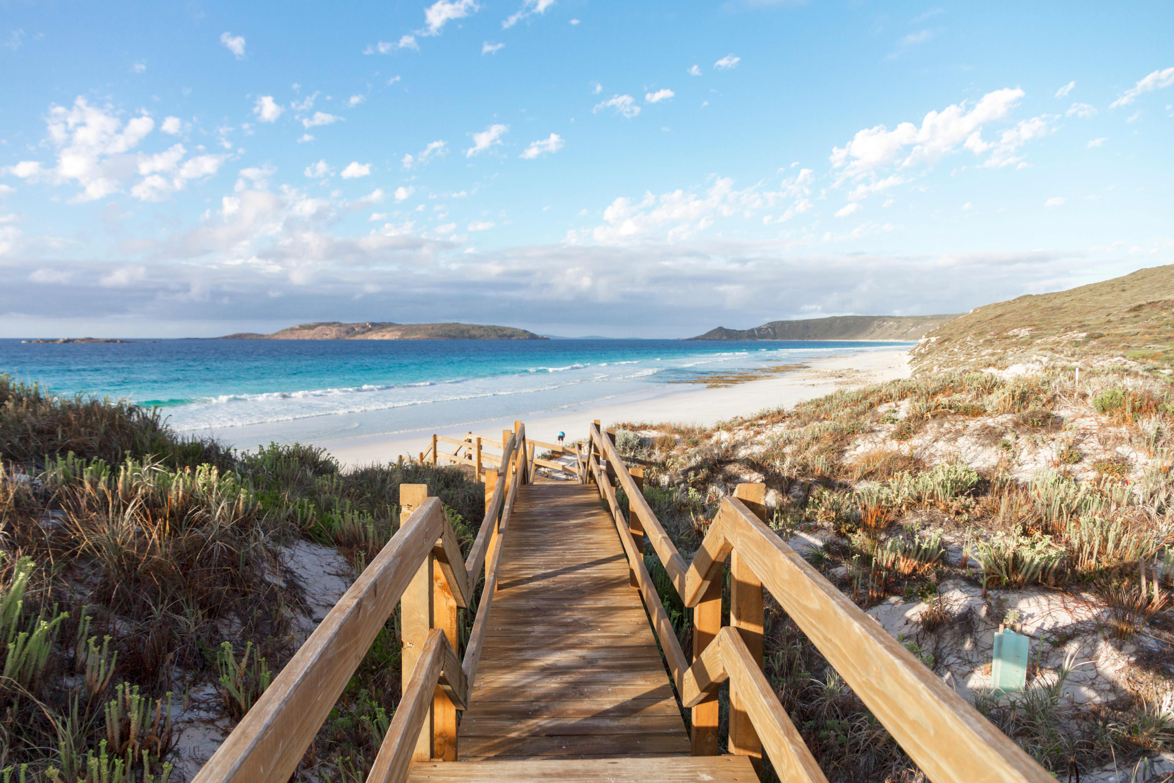 A wooden staircase surrounded by lush vegetation, winding down to a coastal beach with turquoise blue water and white sand.